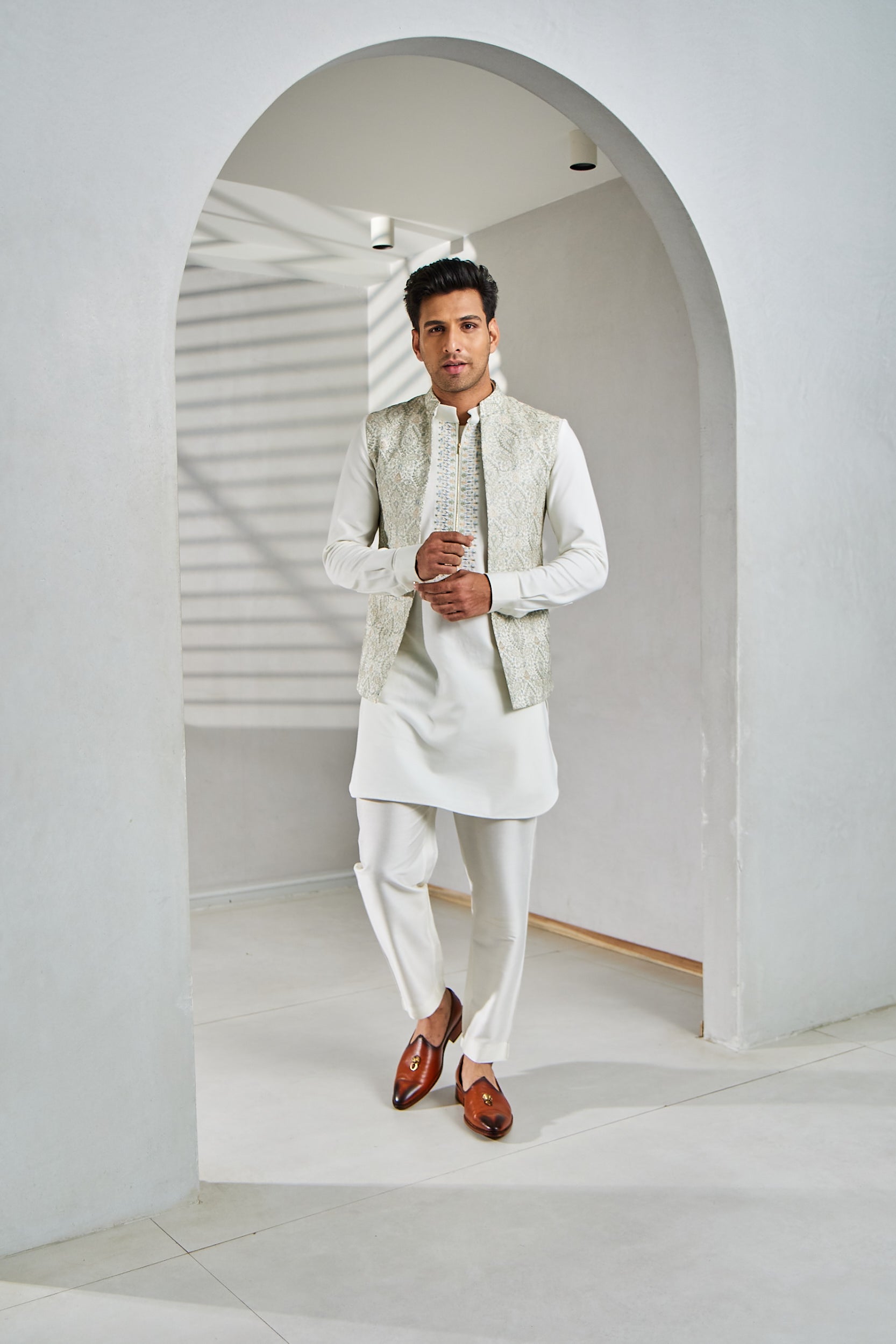 A man poses in a white bundi set which includes a silk bundi, a kurta, and a Ali Ghadi pajama, with resham embroidery.