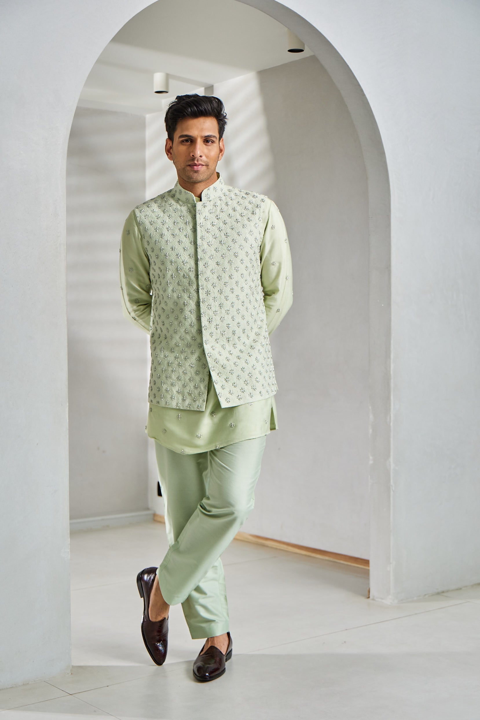 A man poses in a pista green kurta bundi set, featuring a cotton silk kurta and Ali Ghadi paijama, paired with a Banarasi bundi adorned with intricate resham and moti embroidery.