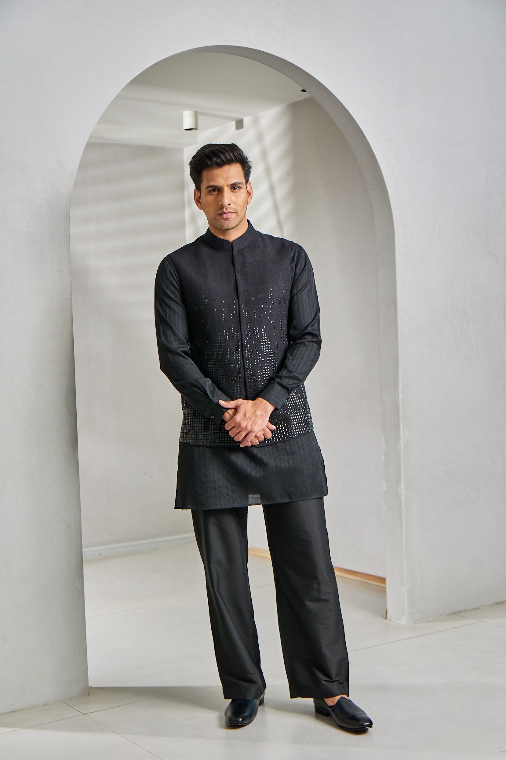 A man poses in a black kurta bundi set featuring a jacquard silk kurta, cotton silk Ali Ghadi paijama, and a matka silk open-style bundi embellished with intricate tikki work at the bottom.