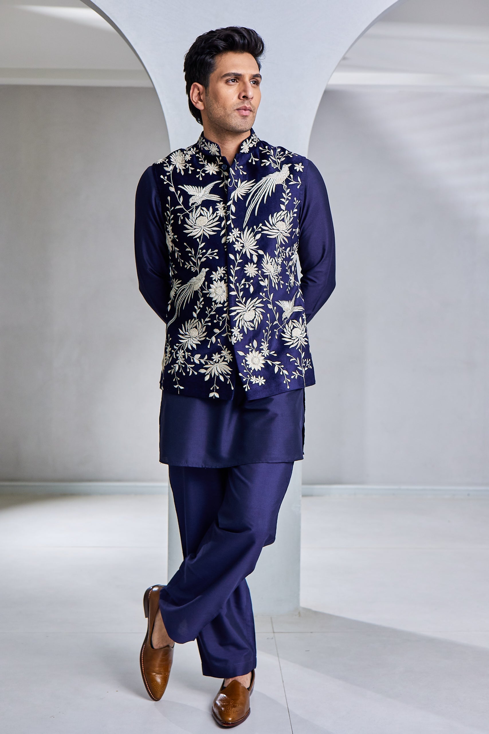 A man poses in a navy blue kurta bundi set, featuring a cotton silk kurta and paijama, paired with a cotton suede bundi adorned with golden resham thread hand embroidery.