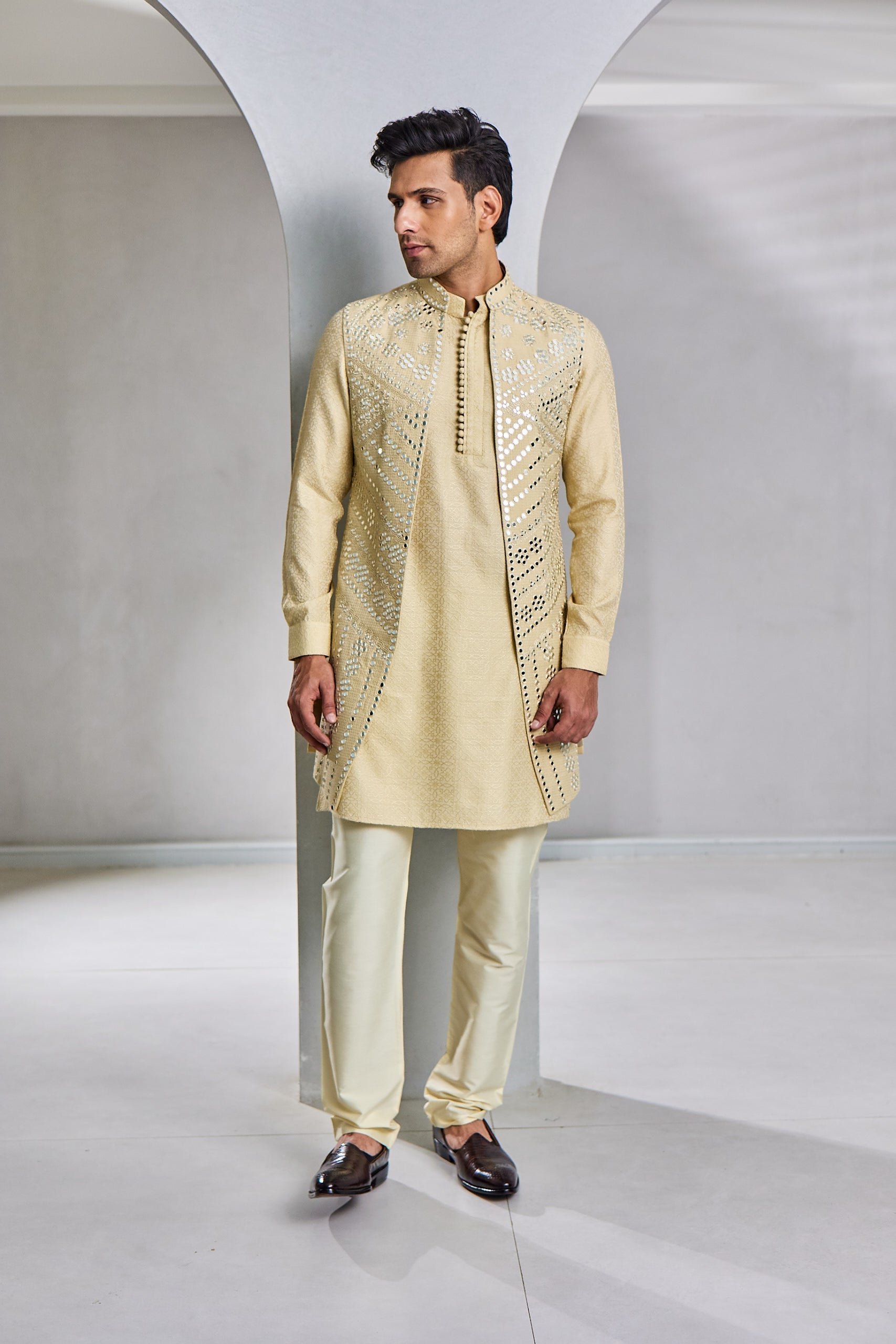 A man poses in a light gold kurta bundi set, featuring a Jacquard silk kurta and Ali Ghadi paijama, paired with a front-open longline bundi adorned with mirror and resham thread work on the front and a plain back.