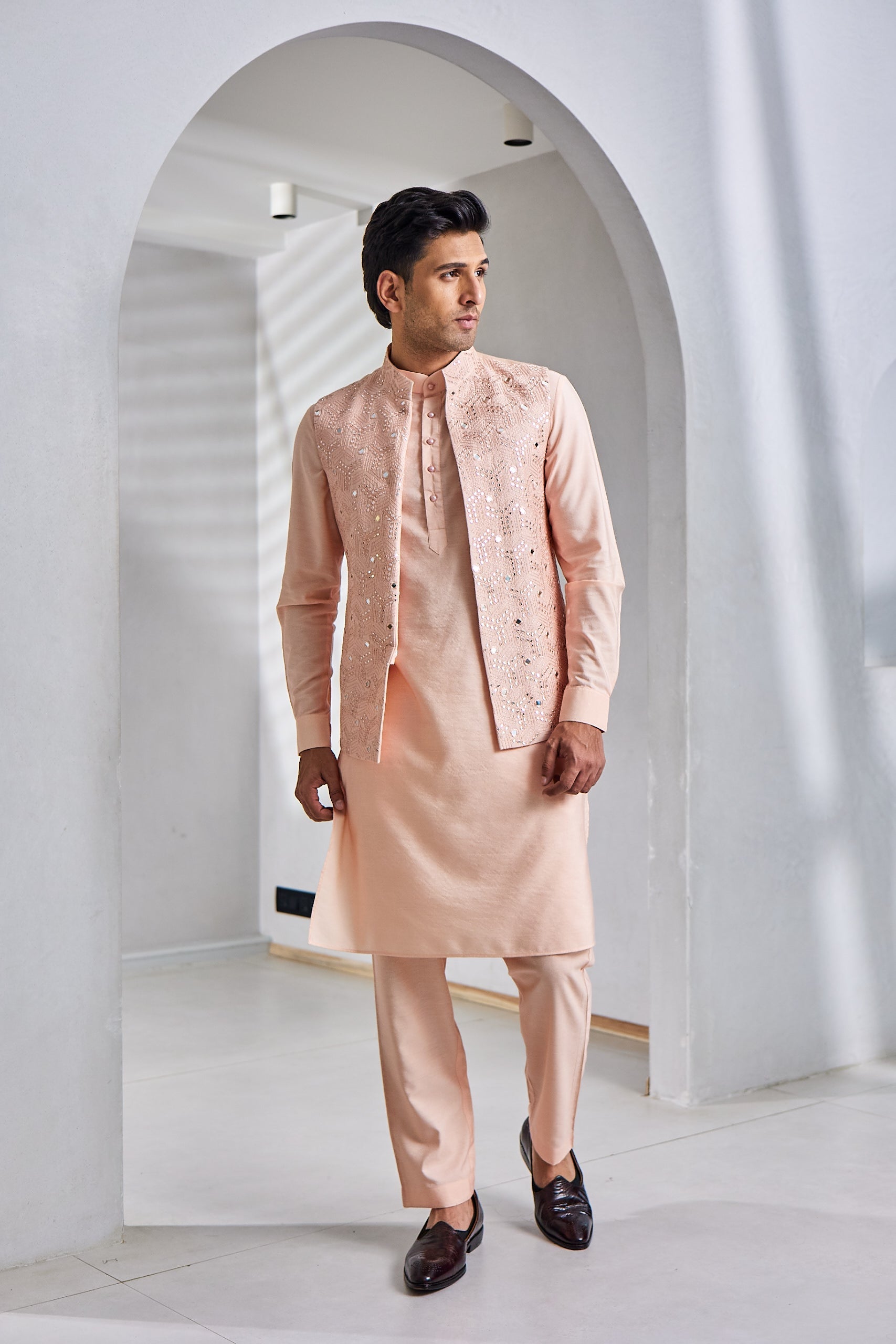 A man poses in a peach kurta bundi set, featuring a Munga silk kurta and bundi with resham thread and mirror sequin work, paired with a cotton silk paijama and finished with pearl half-drilled buttons on the neck.