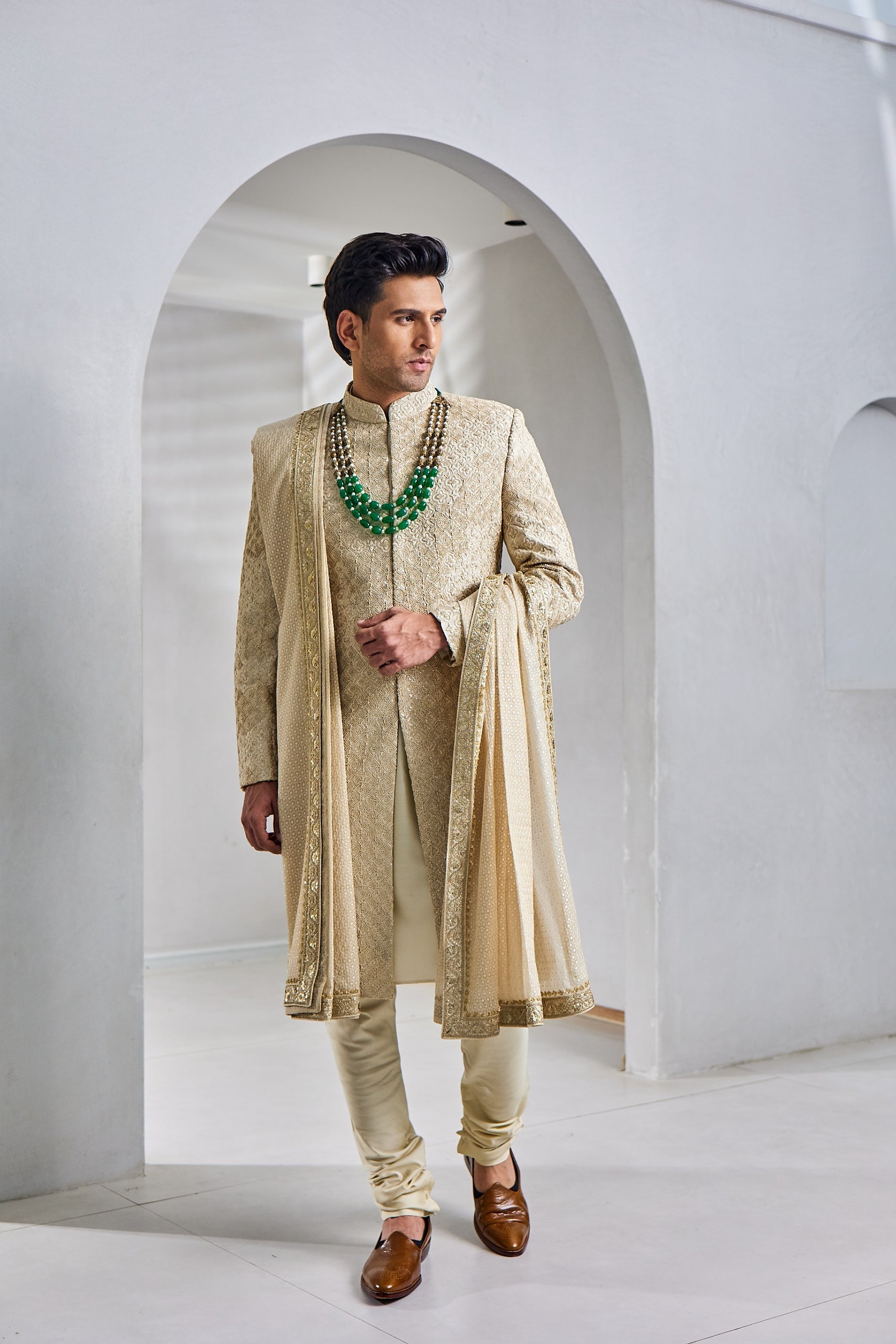 A man poses in a regal beige groom’s sherwani set, featuring a raw silk sherwani with resham and tikki work on the front, sleeves, and back, paired with a plain cotton silk kurta and churidar, and a tissue silk shawl with embroidered borders.