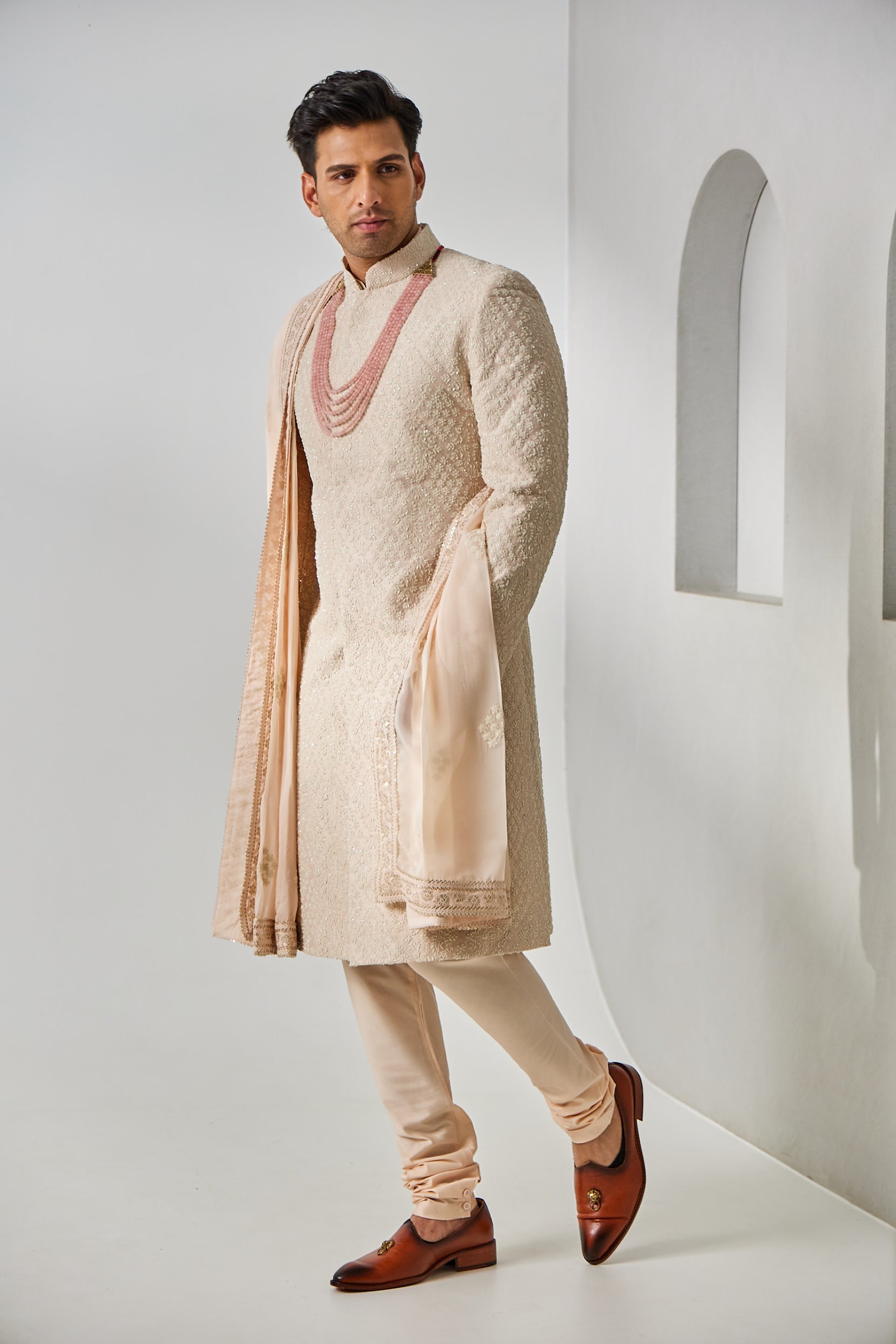 A man poses in a powder pink sherwani set, featuring a raw silk sherwani with resham thread and Kathdana embroidery, paired with a plain cotton silk kurta and churidar, and a tissue silk shawl embellished with matching resham and Kathdana work along the borders.