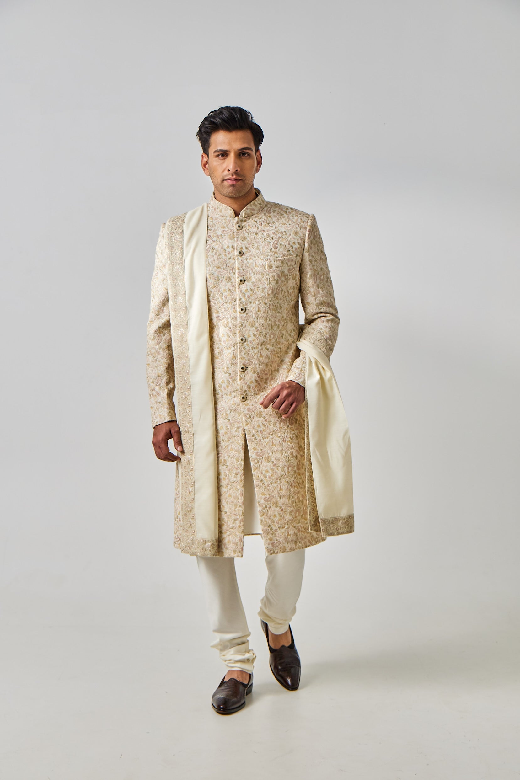 A man poses in a cream sherwani set, featuring a Munga silk sherwani with vibrant multi-colour resham thread embroidery, paired with a plain cotton silk kurta and churidar, and a tissue silk shawl with an embroidered border—perfect for a pastel summer wedding.