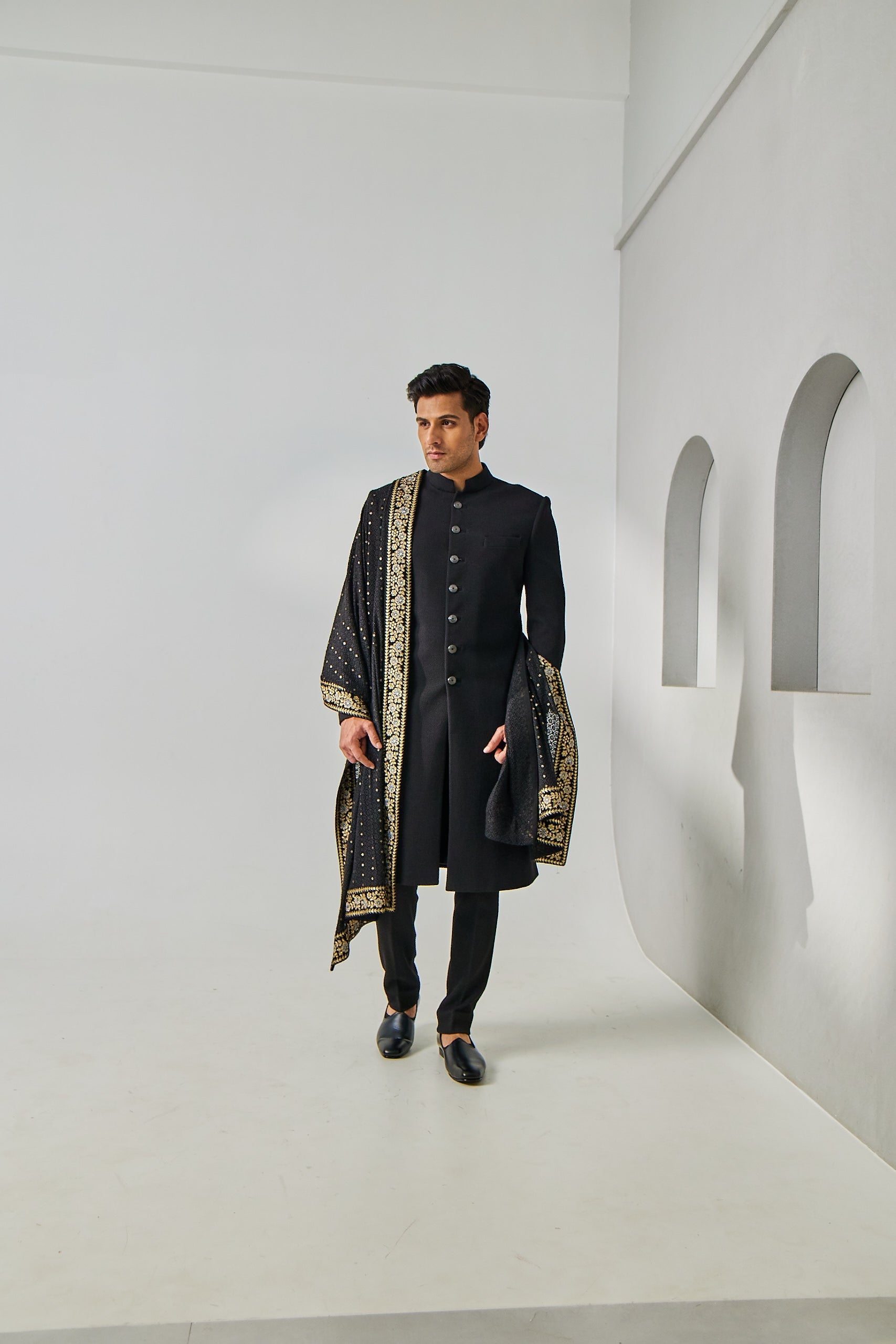 A man poses in a black Nawabi sherwani set, featuring a plain jute silk sherwani and matching trousers, styled with a Lucknowi fabric shawl adorned with resham thread, zari, and tikki embroidery along the borders.
