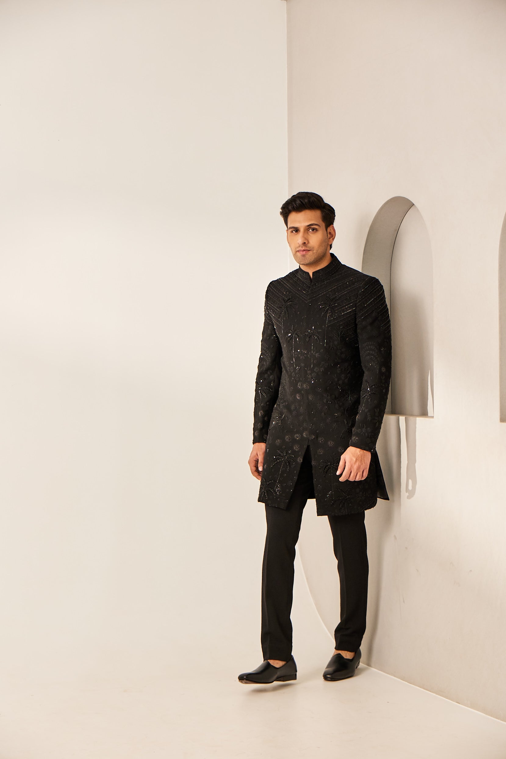 A man poses in a black Indo-Western jacket-style kurta set, crafted from poly wool and paired with trousers, featuring intricate hand embroidery with resham, mirror work, katdana sequins, and zardosi detailing.
