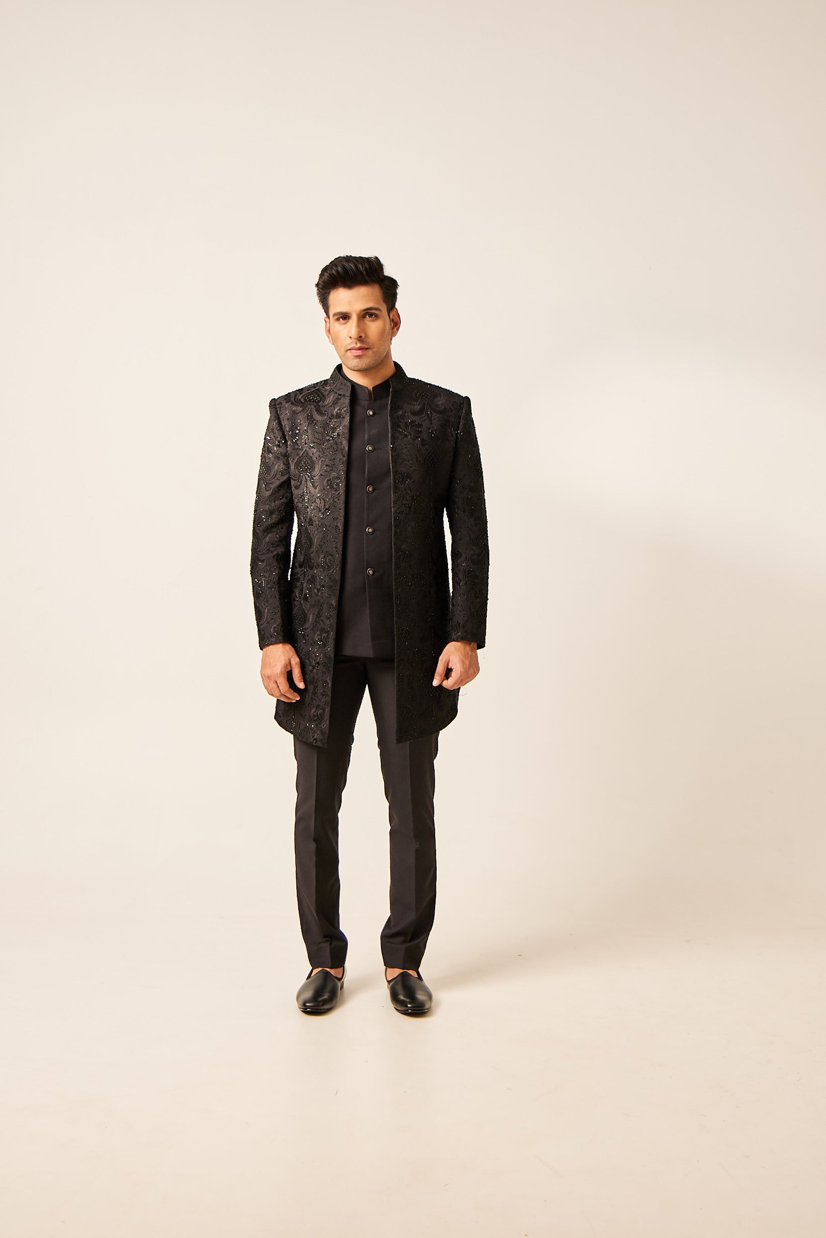 A man poses in a black open-style Indo-Western ensemble in terry wool, featuring hand embroidery with resham, mirror work, katdana sequins, and zardosi. The outfit includes a plain inner bundi and matching trousers.