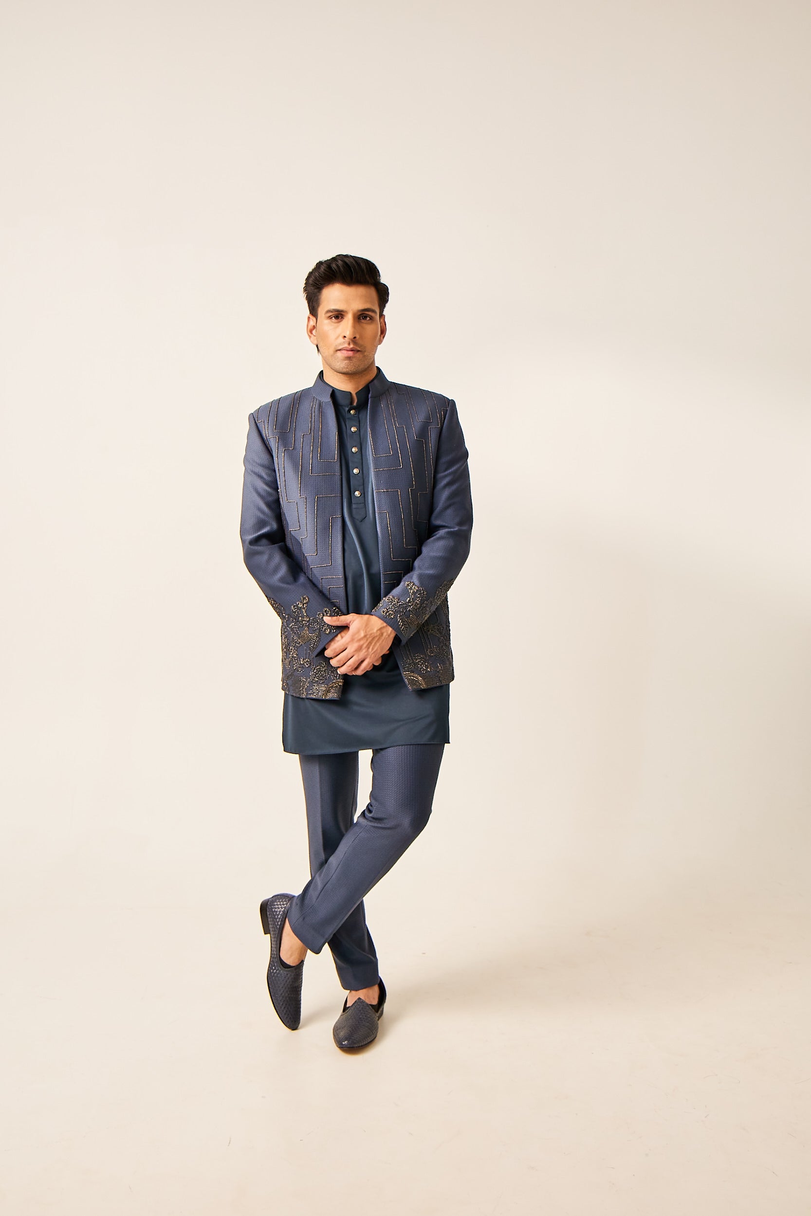 A man models a turquoise Indo-Western ensemble featuring a jacquard jacket with intricate katdana, moti, and antique embroidery. The set includes a plain tencel silk kurta and jacquard trousers, blending traditional elements with modern flair.