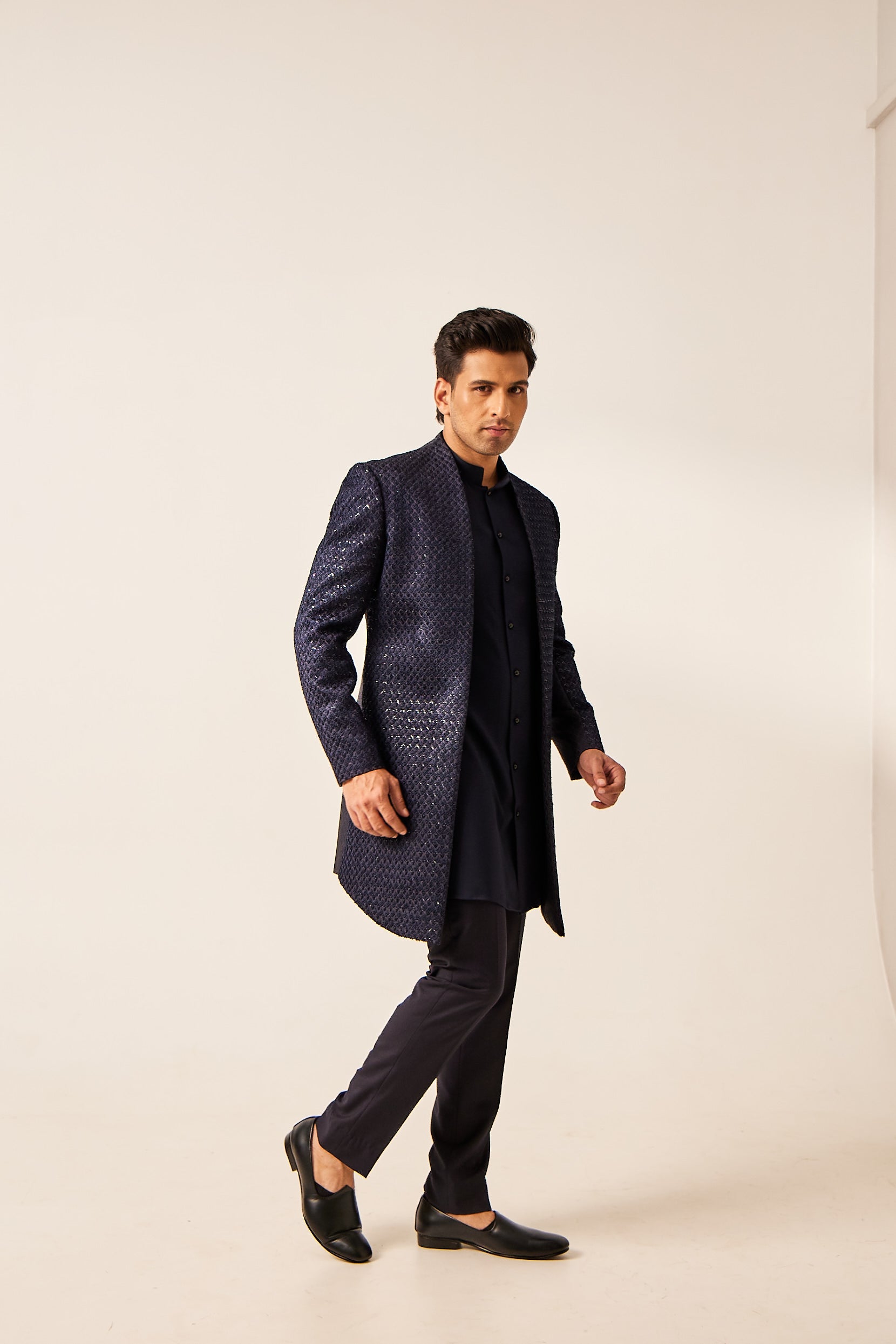A man poses in a blue open-style Indo-Western ensemble featuring a brocade jacket with hand-embroidered resham French knot and tikki work, paired with a plain tencel silk kurta and terry wool trousers.