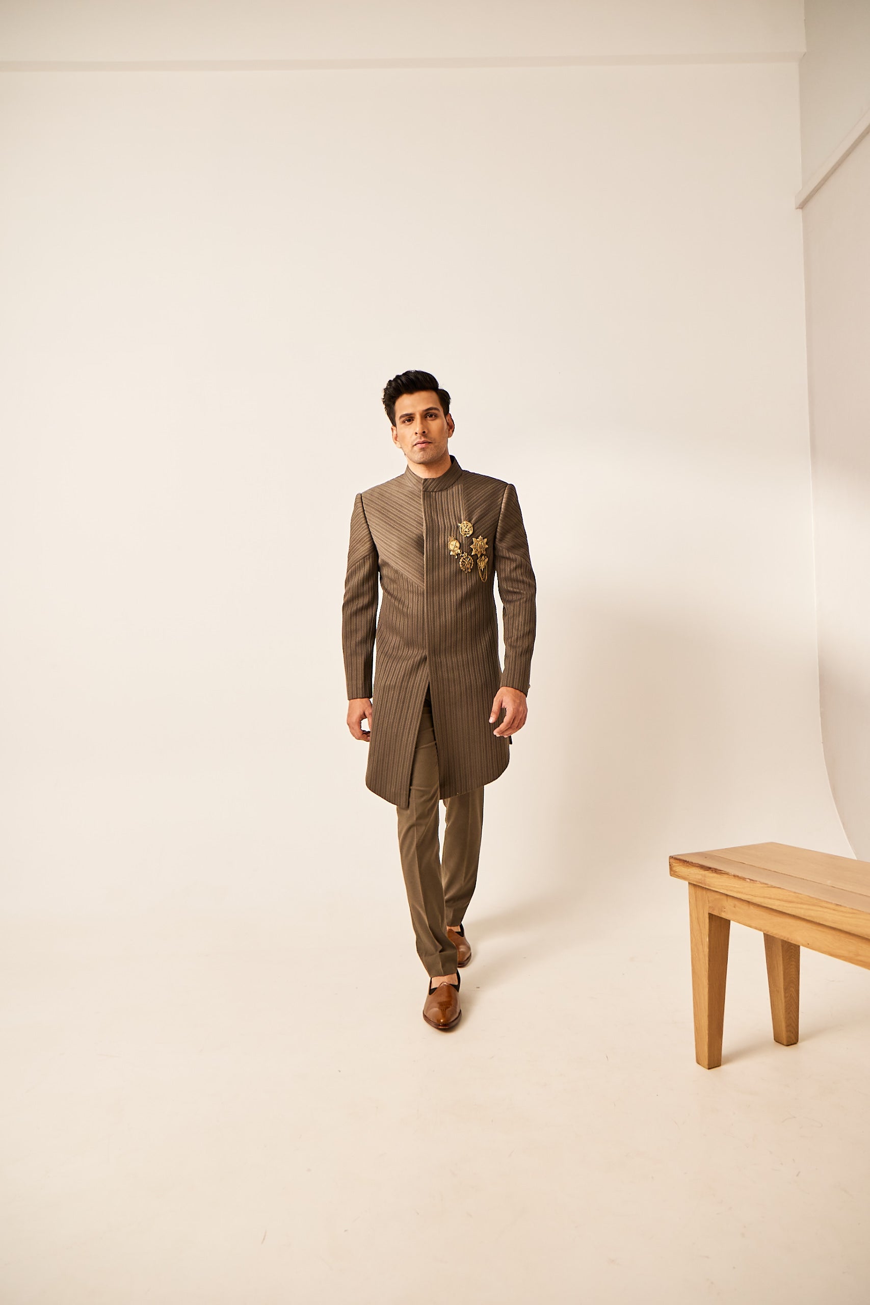 A man poses in an olive green Indo-Western outfit crafted from plain knitted fabric, styled with a statement brooch and paired with matching plain trousers.