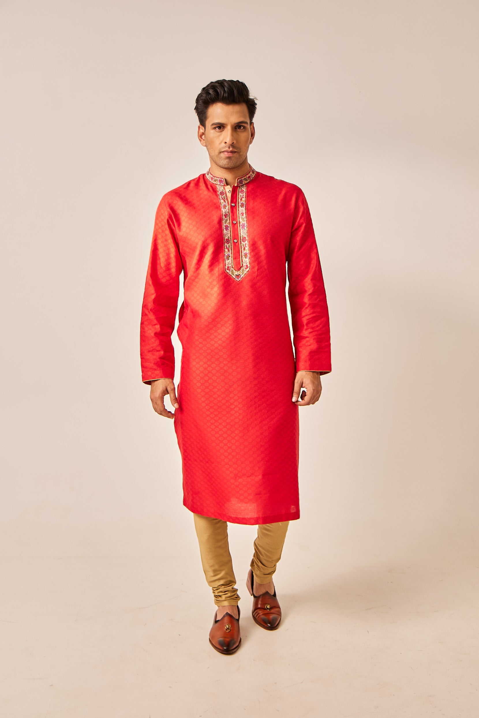 A man poses in a red Banarasi silk kurta adorned with resham Kashmiri thread work on the collar, paired with a red and golden cotton silk churidar for a festive, regal look.
