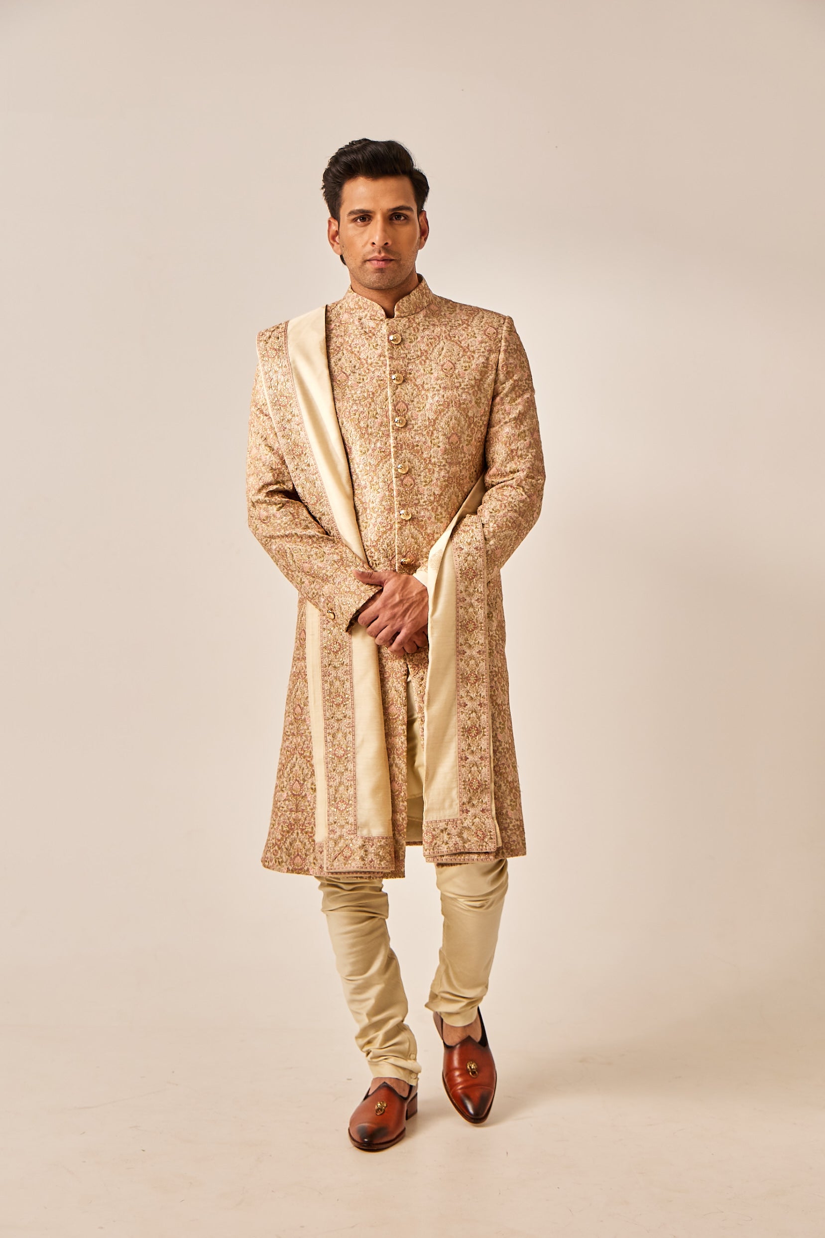 A man poses in a gold Munga silk sherwani adorned with intricate multi-color resham thread work on the front and back, paired with a plain cotton silk kurta, churidar, and a tissue silk shawl featuring an embroidered border.