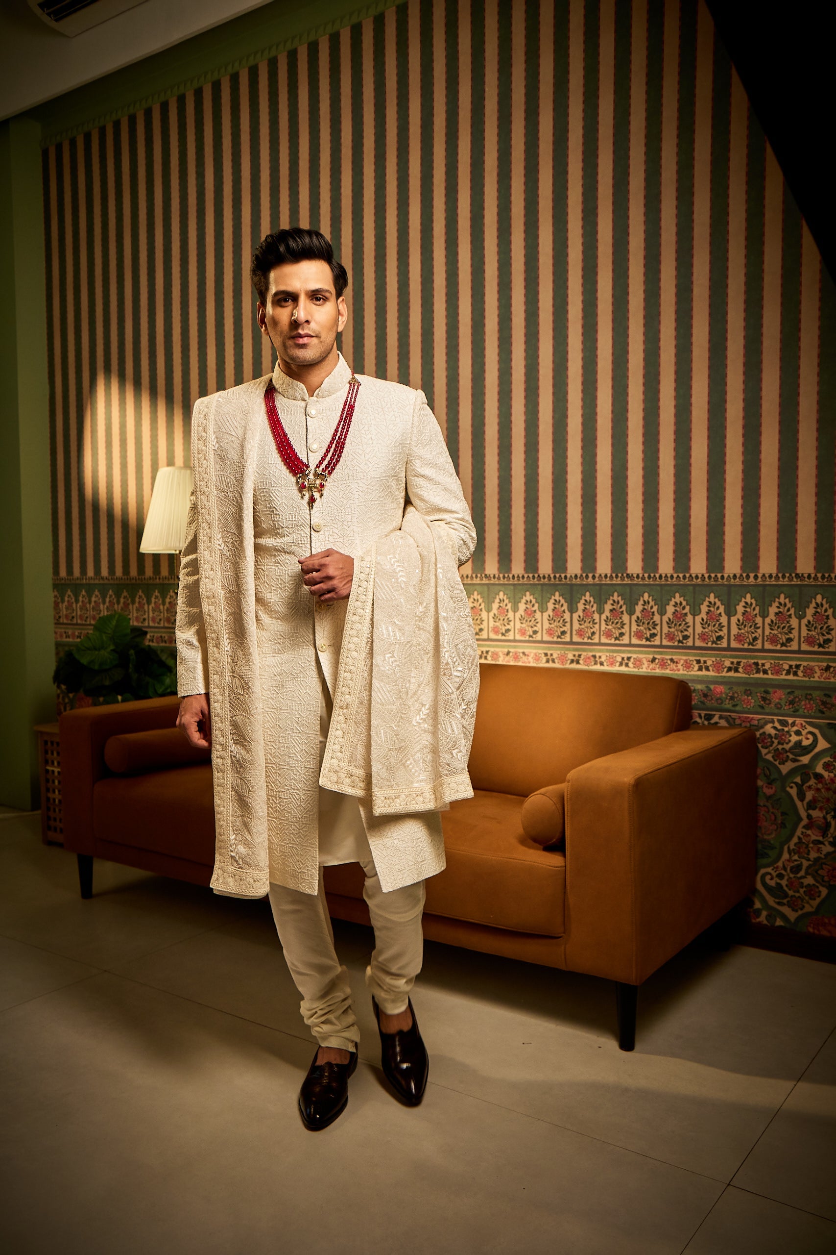 A man poses in an off-white raw silk sherwani embellished with resham thread, moti, and tikki work, paired with a cotton silk kurta-churidar and a net shawl for an elegant ceremonial look.