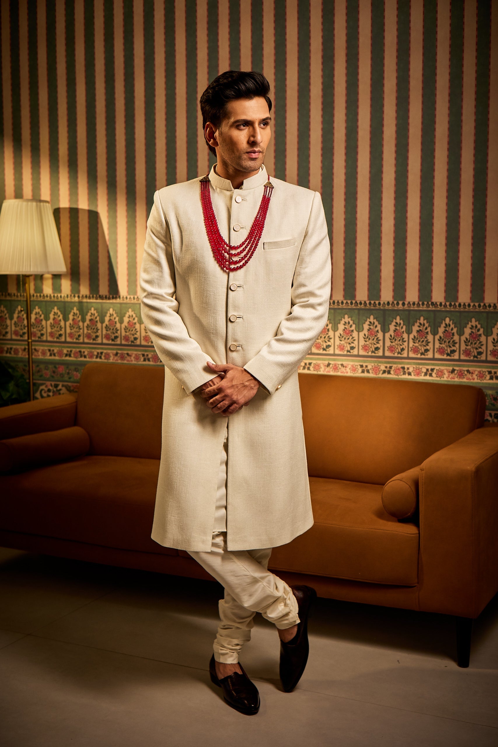 A man poses in a white Jude silk sherwani with a clean, plain design, paired with a cotton silk kurta and churidar for a classic, wedding-ready look.