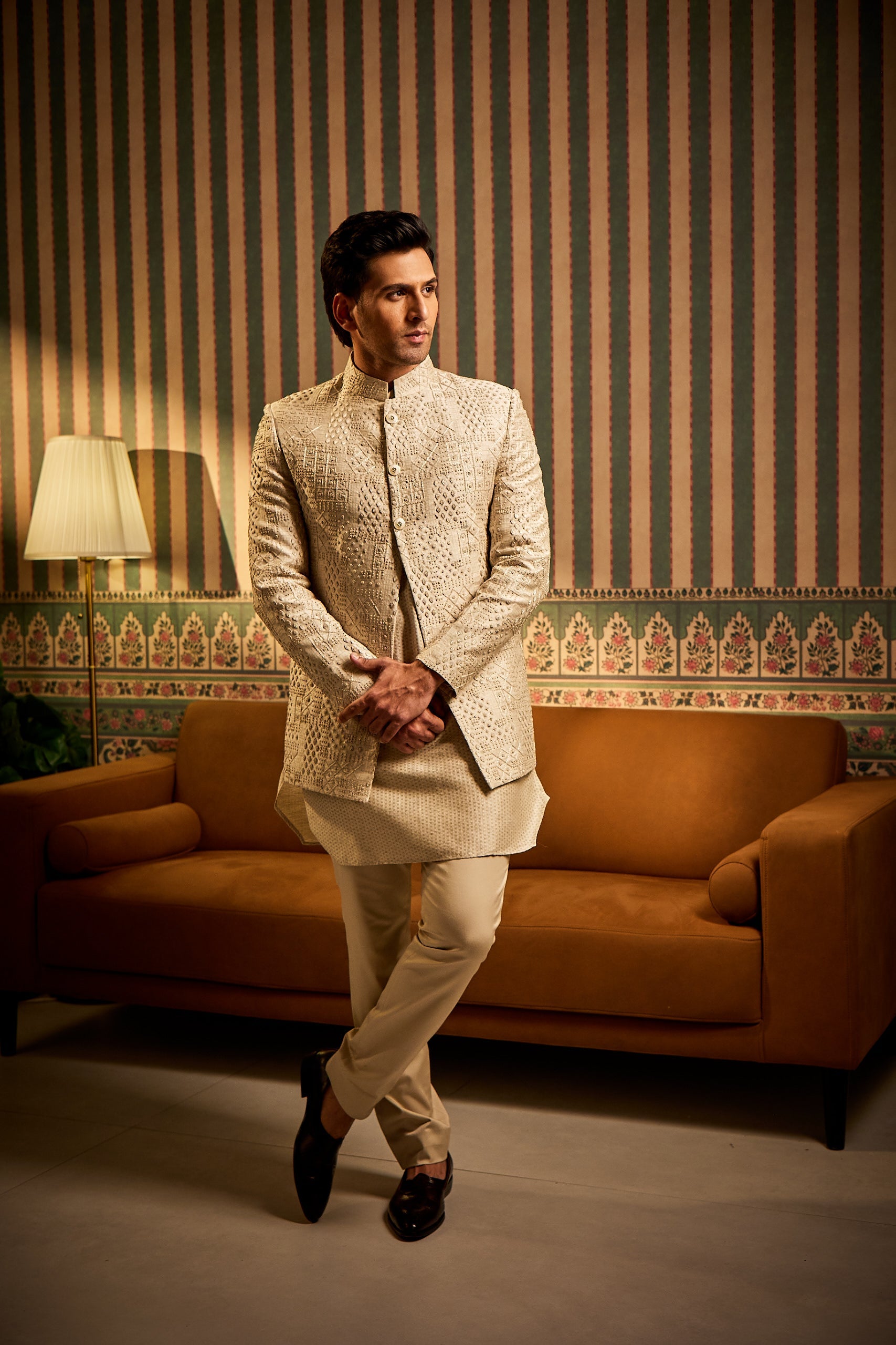 A man poses in a beige Banarasi silk Indo-Western ensemble featuring intricate resham thread French knot embroidery, paired with a silk jacquard kurta and matching silk trousers for a refined fusion look.