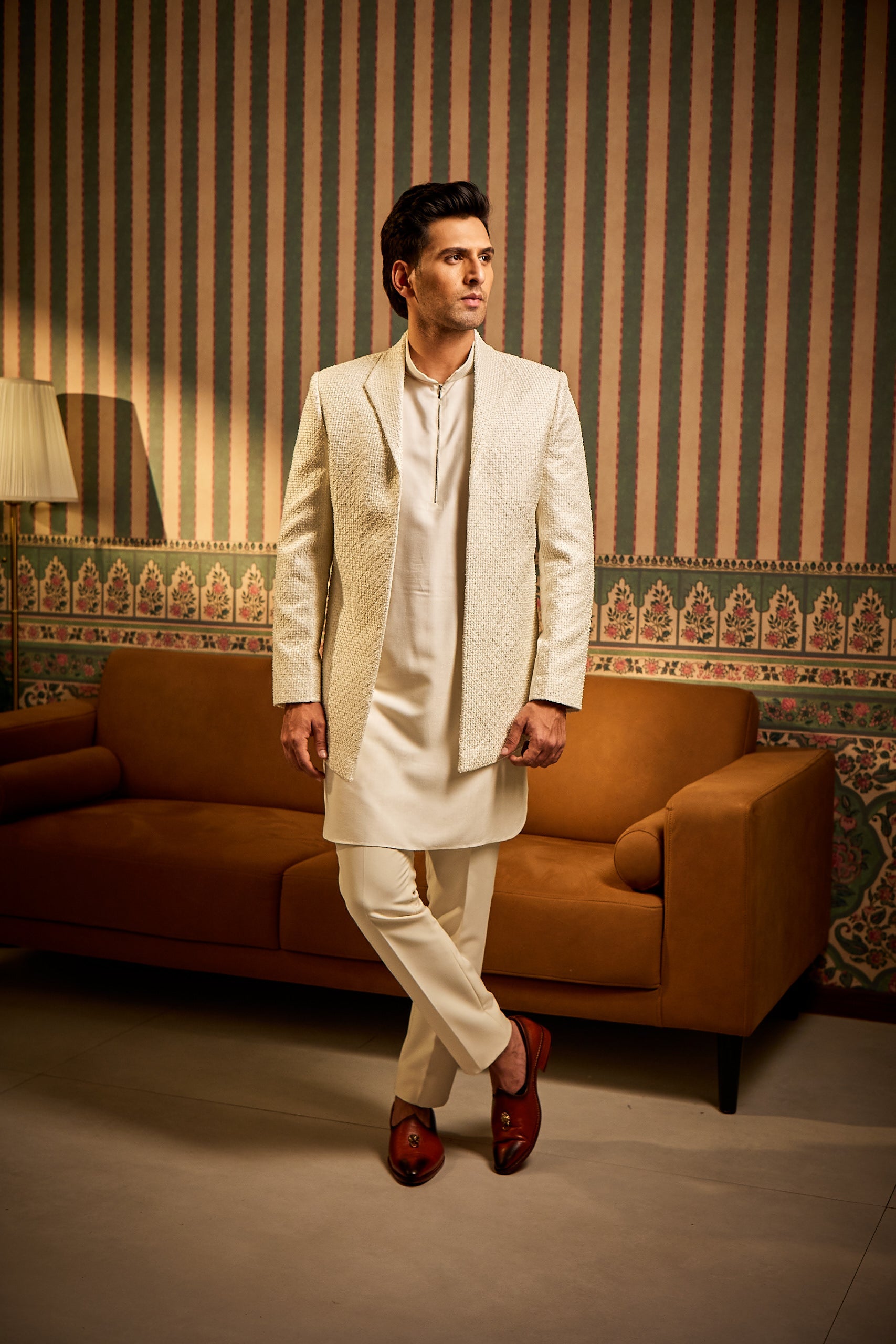 A man poses in an off-white Indo-Western outfit featuring a brocade jacket with resham thread, moti work, and French knot embroidery, layered over a tencel silk kurta and paired with cotton trousers for a graceful fusion look.