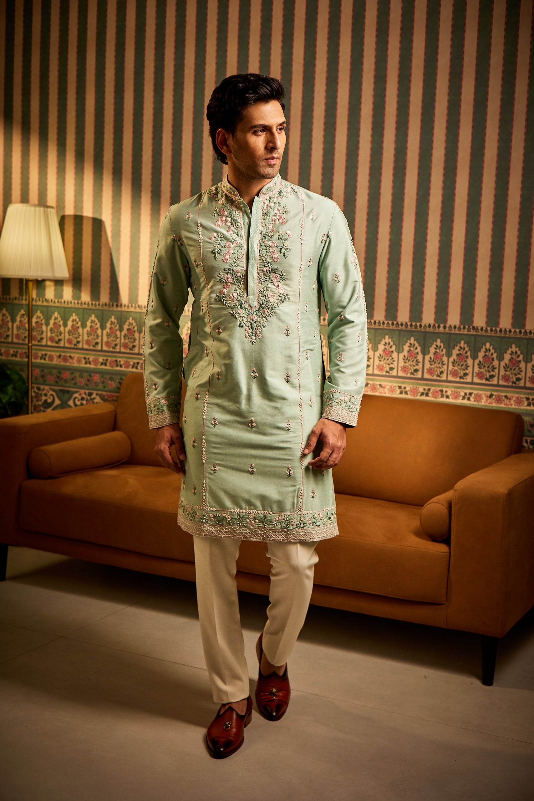 A man poses in a turquoise cotton silk kurta with hand-embroidered multi-color resham thread work on the front and sleeves, paired with a matching Ali Ghadi trouser for a vibrant and graceful look.