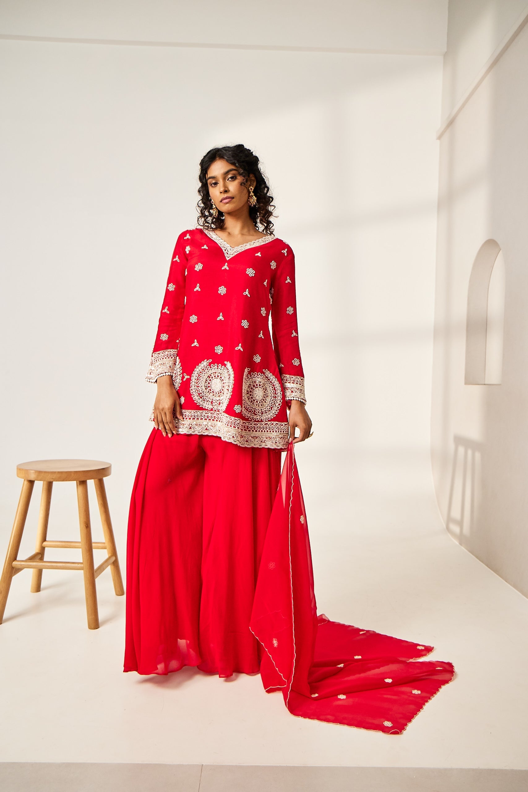 A woman poses in a red co-ord set which includes a silk V-neck kurta, an organza dupatta, and georgette sharara pants.
