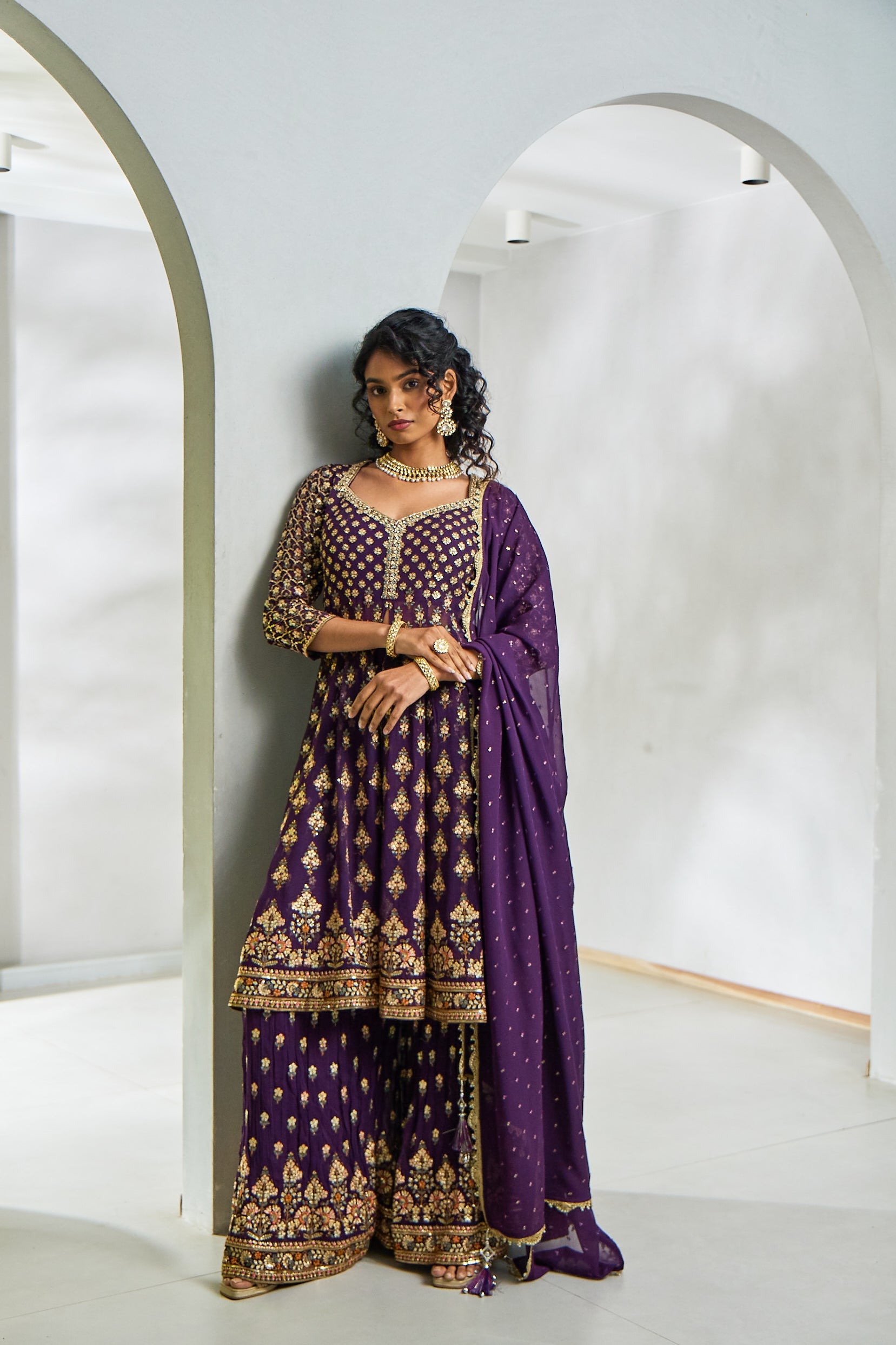 A woman poses in a wine georgette sharara set featuring a short open-style frock with resham thread work and sequin embroidery, paired with flowy sharara pants and an embroidered soft net dupatta.