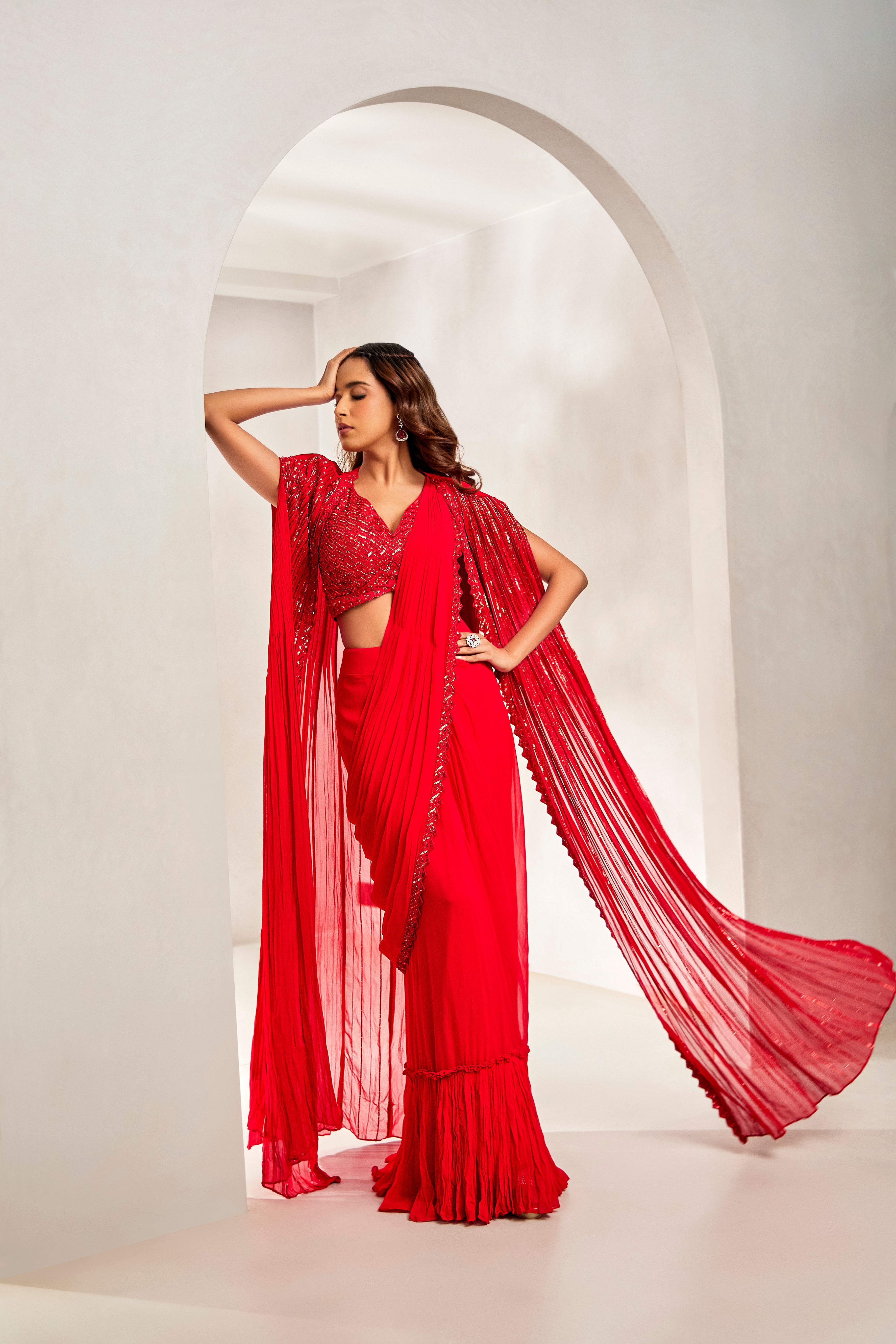 A woman drapes a red pre-draped georgette saree, styled with an embroidered blouse and sheer cape. The ensemble is detailed with Katdhana, sequins, and tikki work for a modern festive flair.