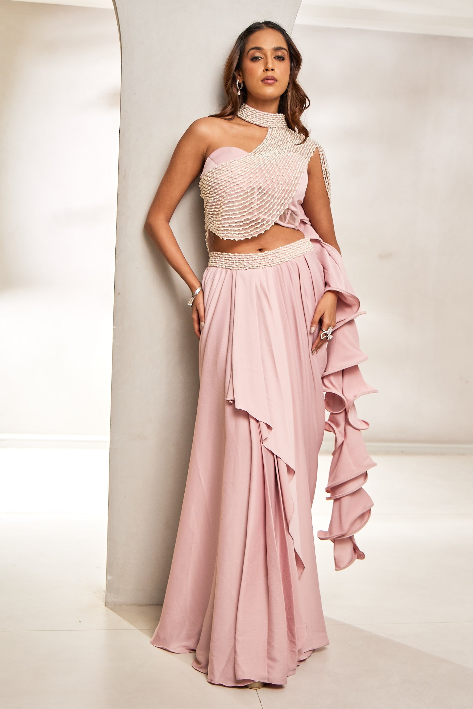 A woman exudes modern elegance in a pink one-shoulder net top and cocktail satin draped skirt. The top is hand-embroidered with delicate pearls and stones, paired with a ruffle dupattaâ€â€perfect for a chic sangeet soirée.