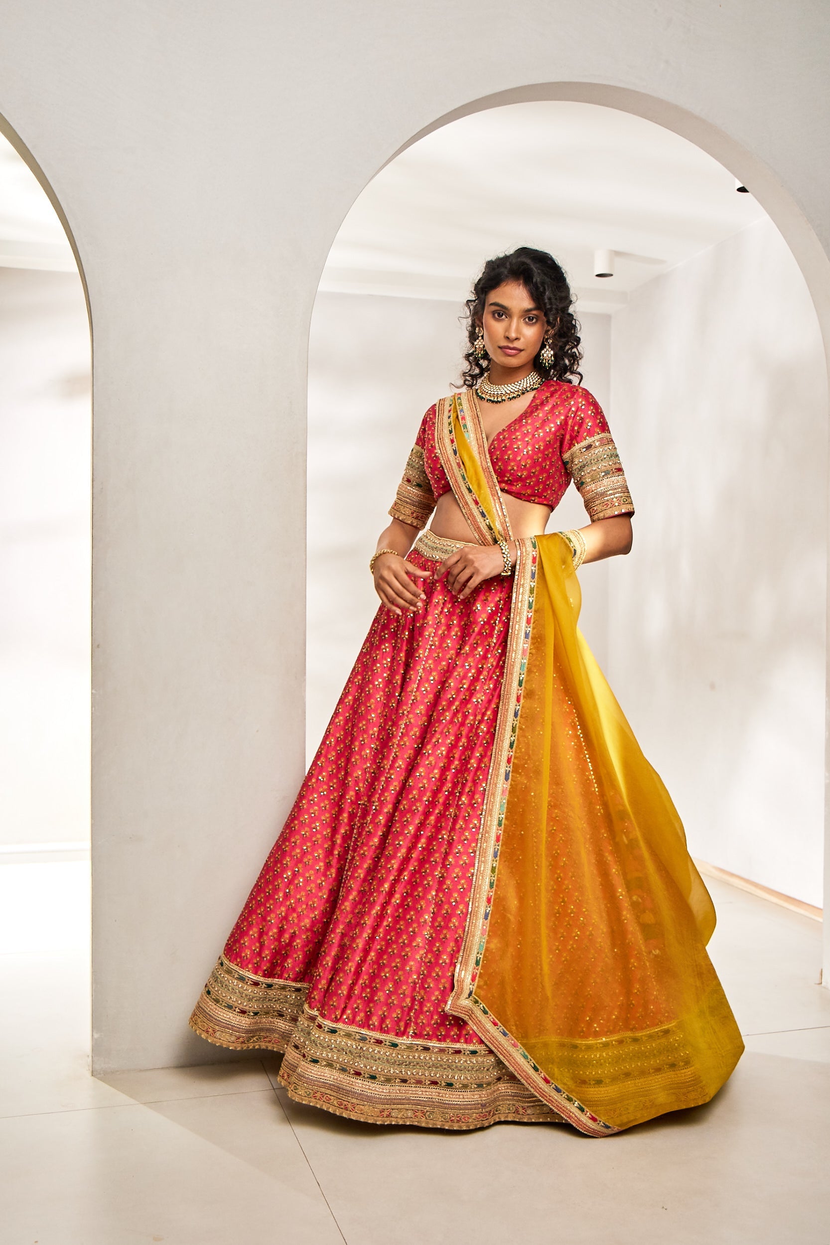 A woman shines in a vibrant pink and yellow silk ghagra choli set, paired with a sheer organza dupatta. The ensemble features multicolour thread, sequin, dori, and bead embroideryâ€â€an eye-catching choice for sangeet celebrations.