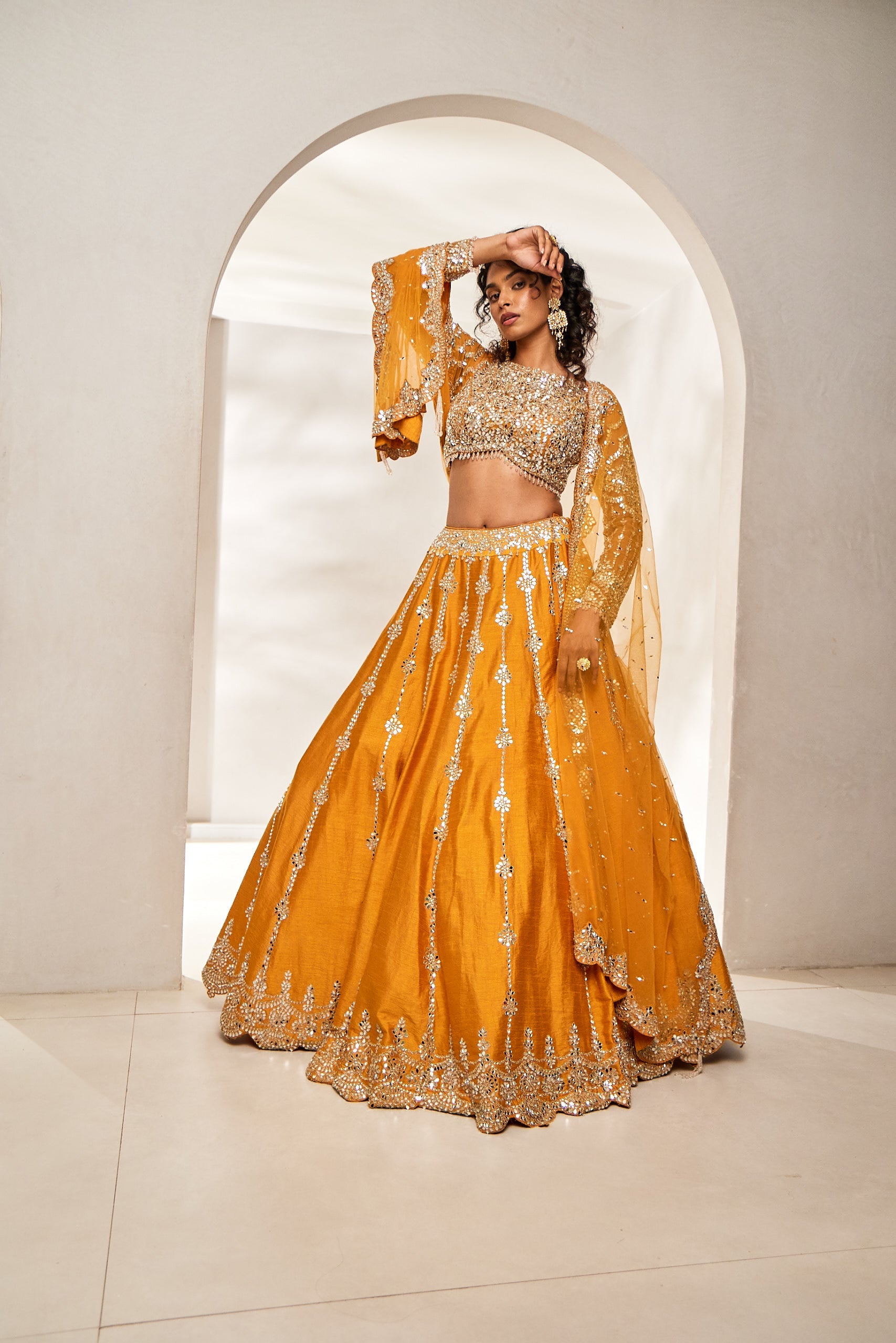 A woman radiates elegance in a marigold silk ghagra choli set, paired with a net blouse and dupatta. The ensemble sparkles with intricate mirror, stone, and dori workâ€â€made for the spotlight of a sangeet night.