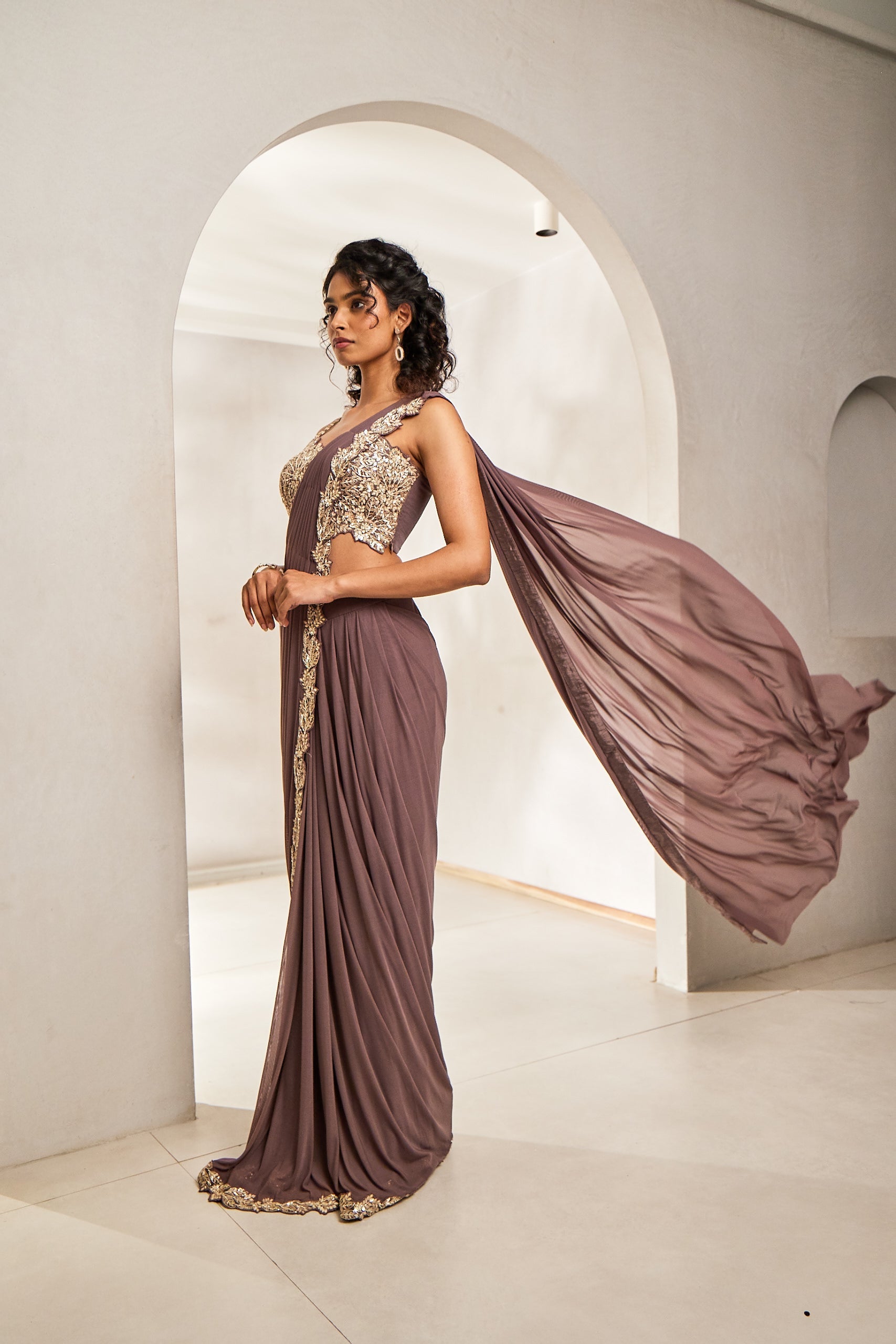 A woman dons a mauve pre-draped lycra net saree paired with an organza blouse. The blouse and saree border are embellished with Katdhana, sequins, stones, and hand-dyed bead embroidery, creating a refined Indo-western look.
