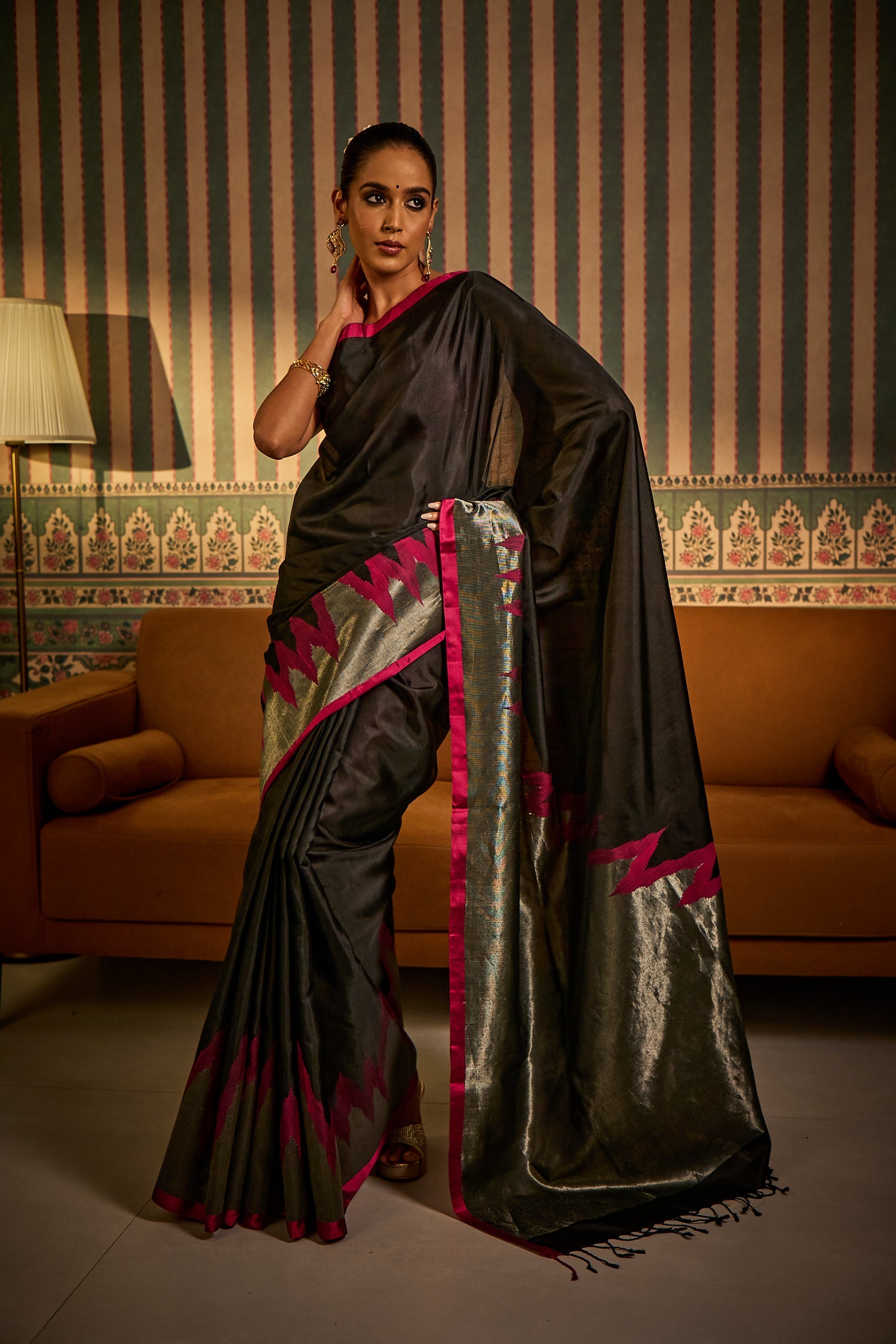 A woman poses in a black south silk saree, showcasing handwoven detailing and a striking golden and pink contrast pallu, styled with a matching blouse fabric and petticoat.