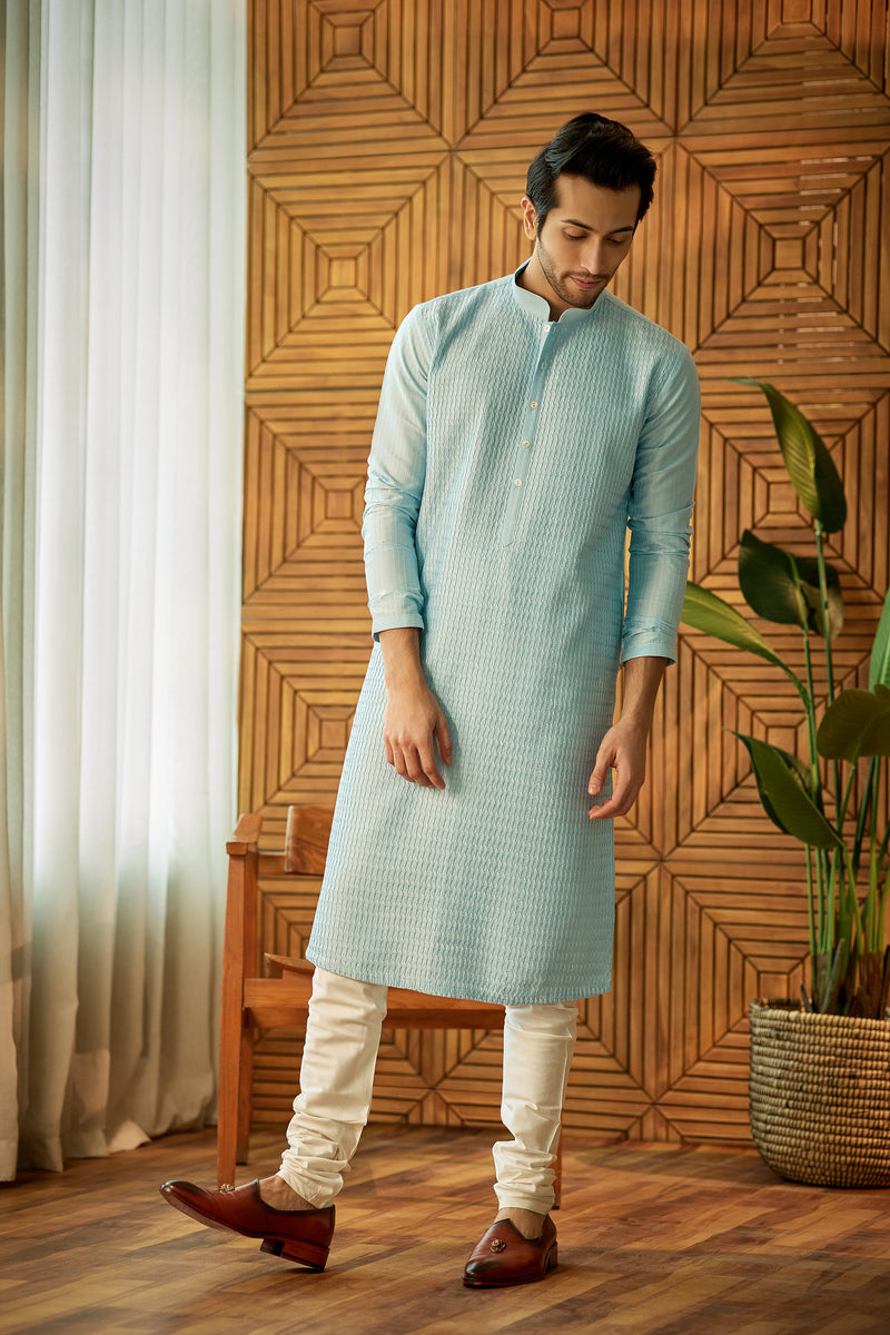 A man in an aqua cotton silk kurta set from BenzerWorld ethnicwear store poses in front of a wooden chair and wall.