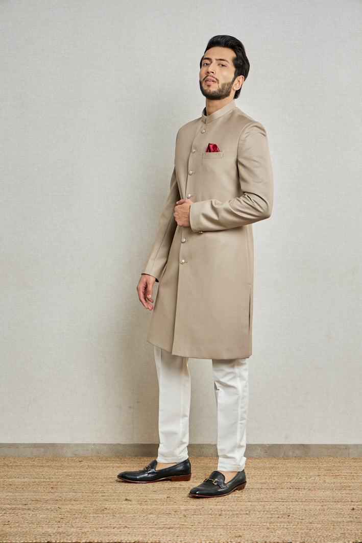 A man in a beige jute silk nawani sherwani from BenzerWorld ethnicwear store poses in front of a wall.