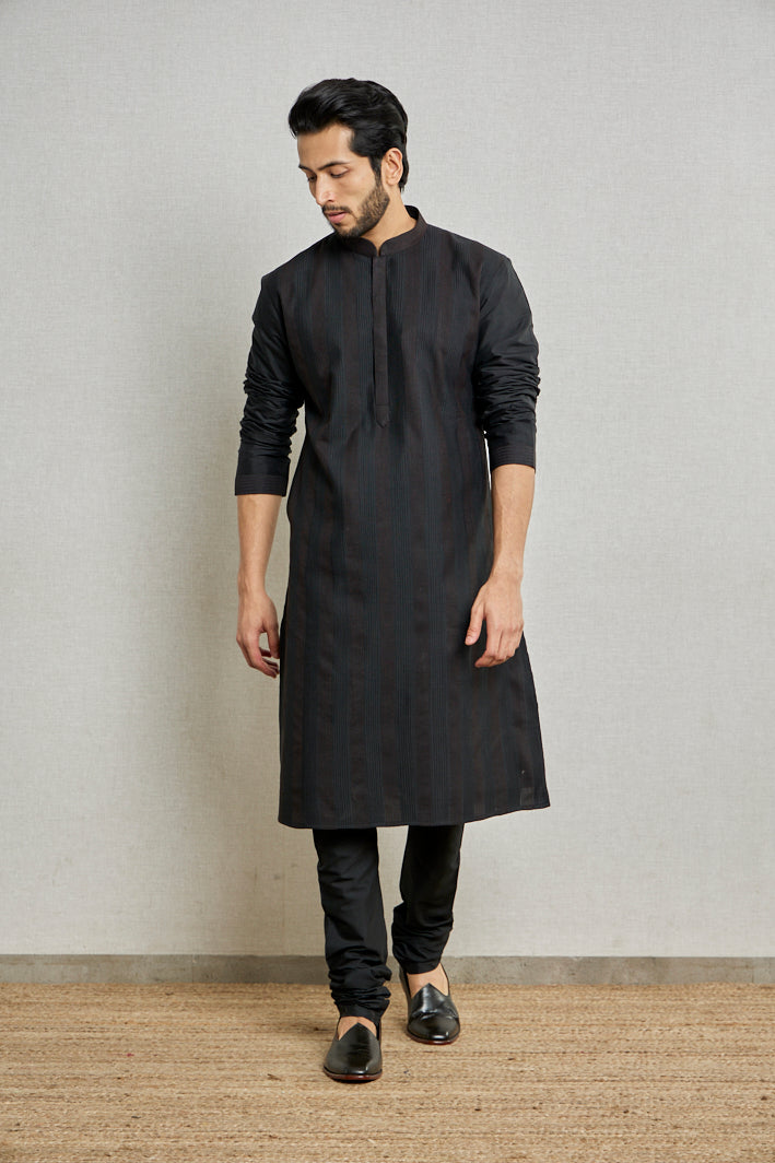 A man poses in a black cotton silk kurta set from BenzerWorld ethnicwear store while standing in front of a wall.