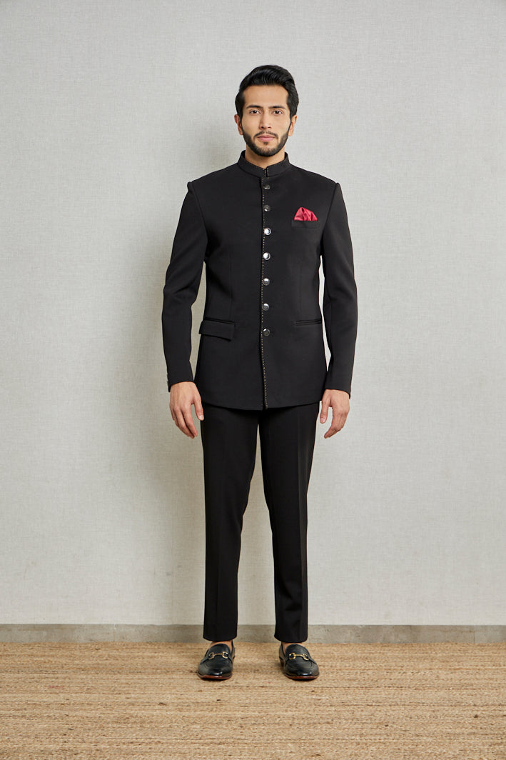 A man poses in a black jute bandgala from BenzerWorld ethnicwear store while standing in front of a wall.