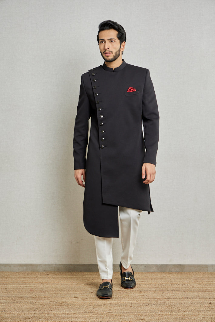 A man in a black jute silk asymmetric Indo-Western suit from BenzerWorld ethnicwear store poses in front of a wall.
