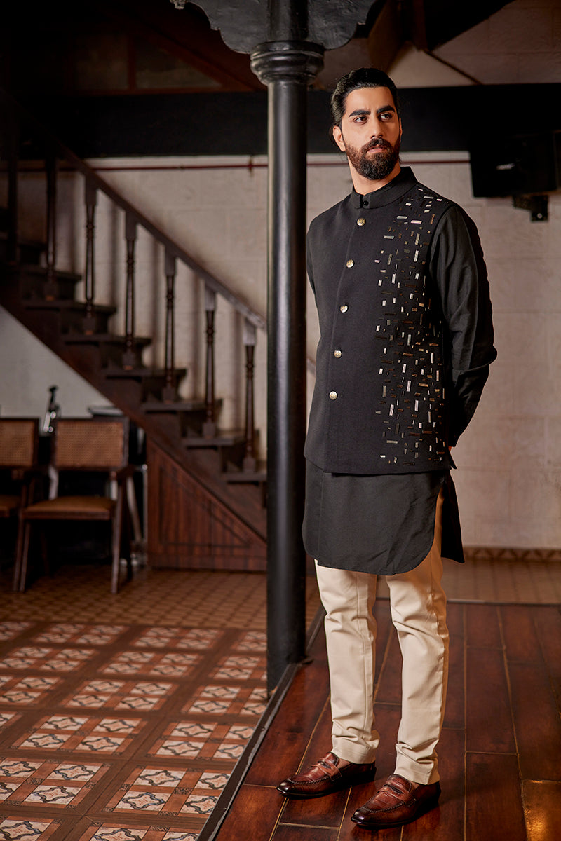 A man in a black matka silk bundi set from BenzerWorld ethnicwear store poses in front of a pole.