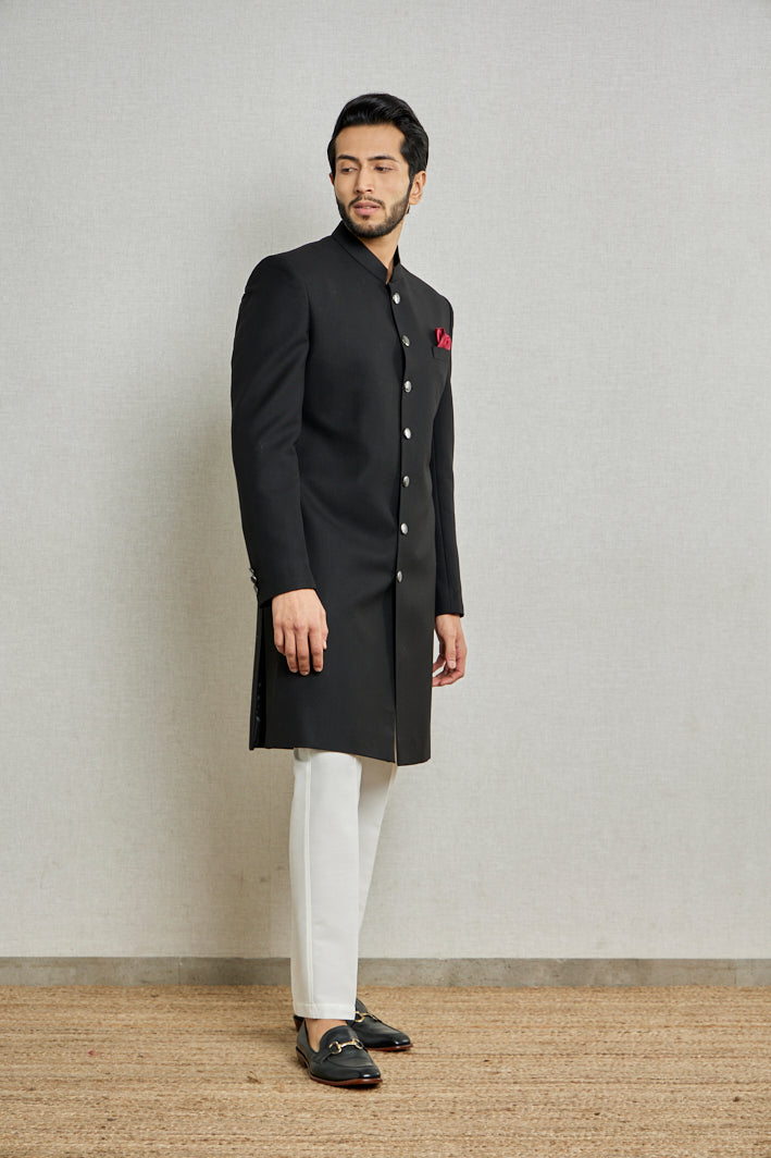 A man poses in a black jute Indo-Western suit from BenzerWorld ethnicwear while standing in front of a wall.