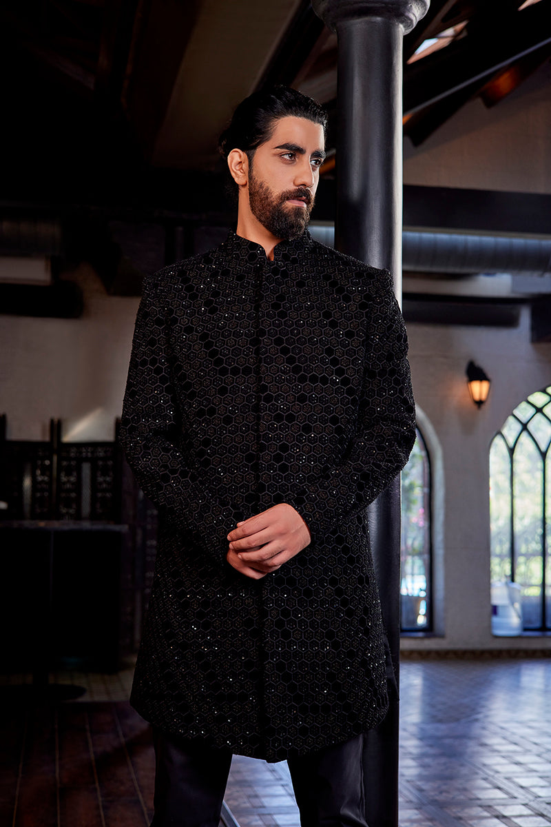 A man in a black velvet Indo-Western suit from BenzerWorld ethnicwear store poses in front of a pole.