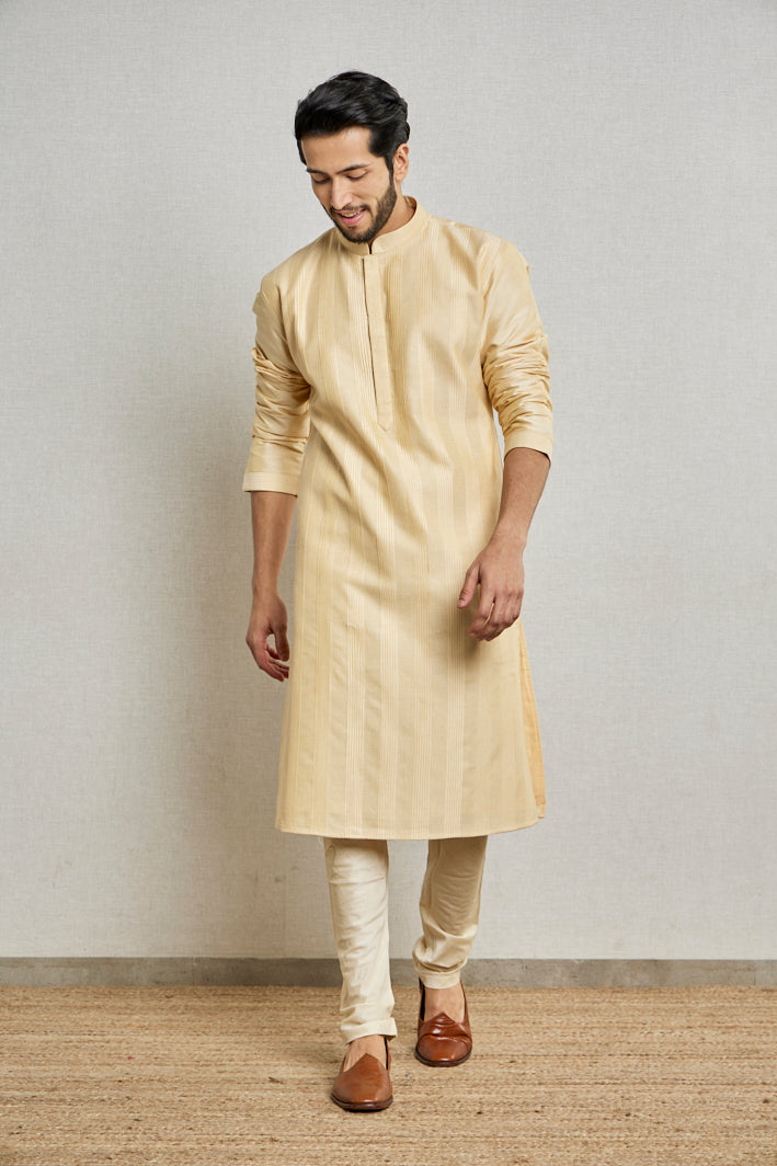 A man poses in a cream cotton silk kurta set from BenzerWorld ethnicwear store while standing in front of a wall.