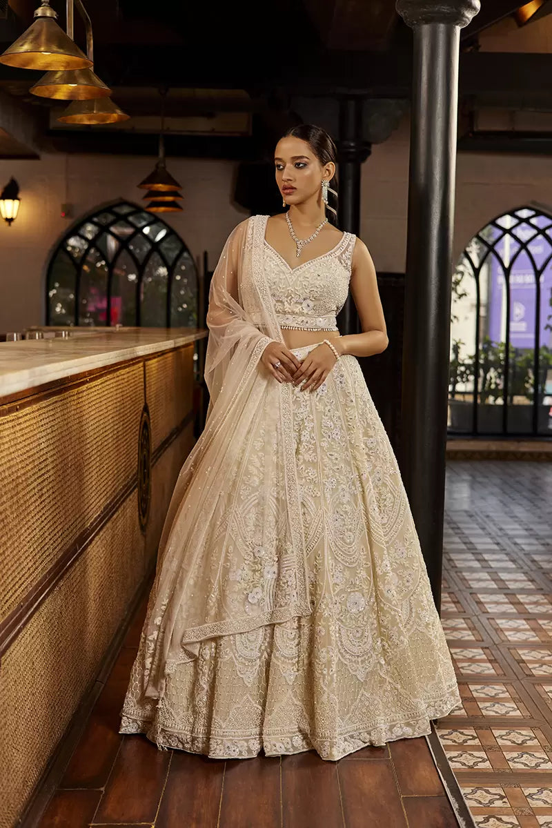A woman poses in a cream organza bridal lehenga from BenzerWorld ethnicwear store, while standing near a column.