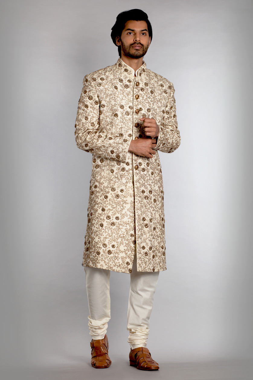 A man poses in a cream matka silk groom's sherwani from BenzerWorld ethnicwear store.