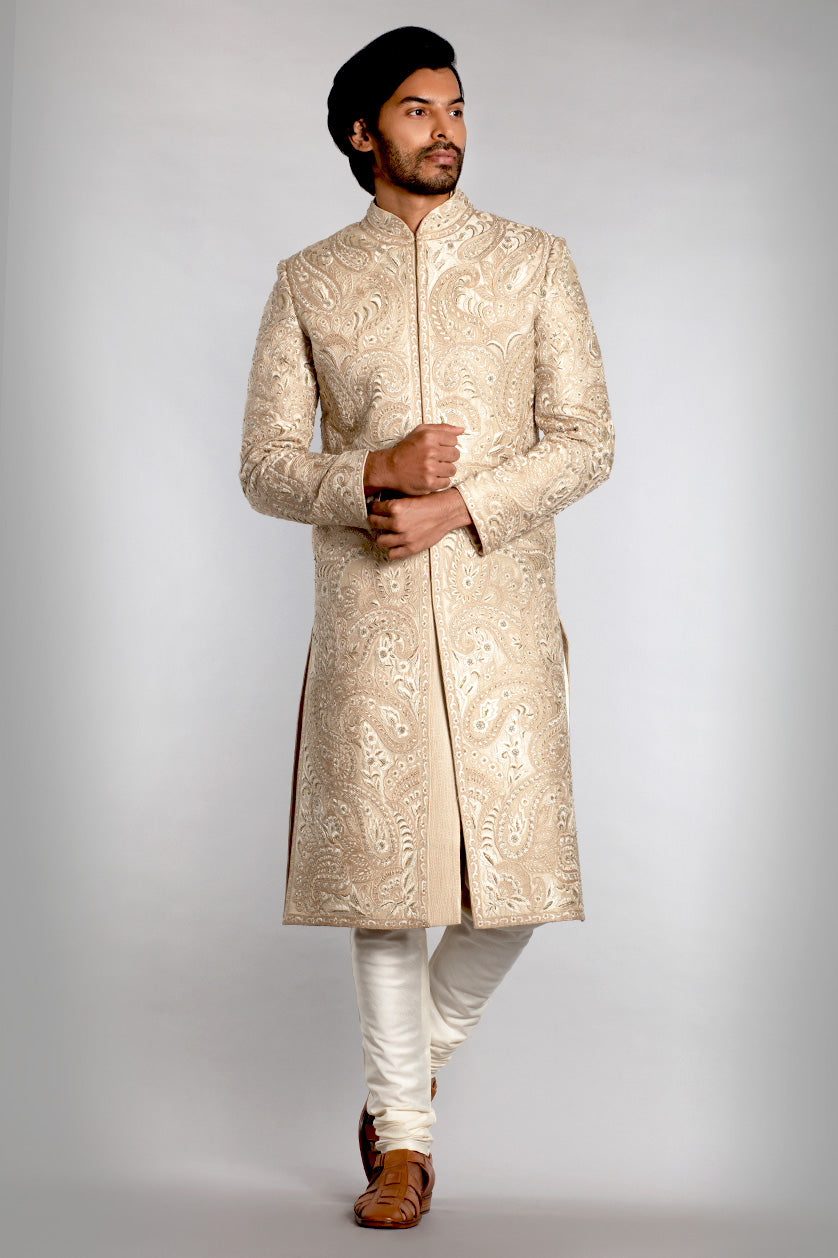 A man poses in a cream matka silk groom's sherwani from BenzerWorld ethnicwear store.