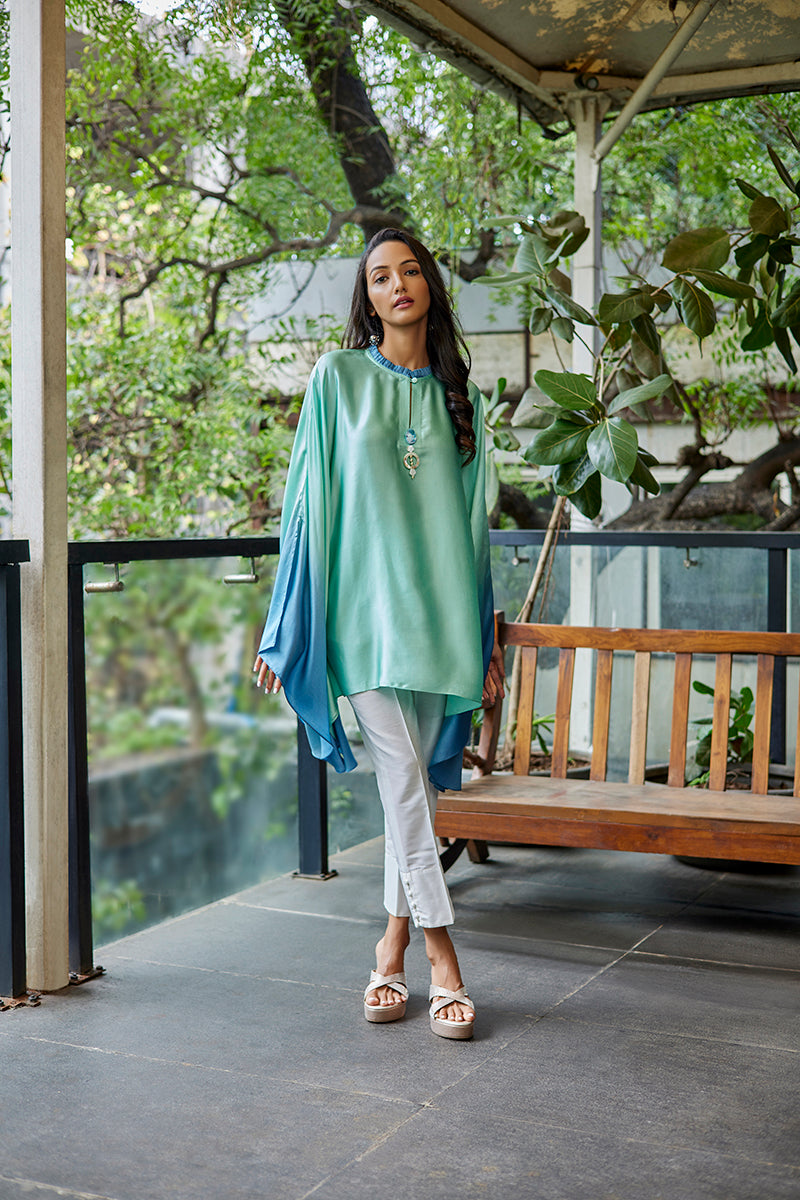 A woman poses in a green-blue satin silk short kurti from BenzerWorld ethnicwear store, while standing near a bench.