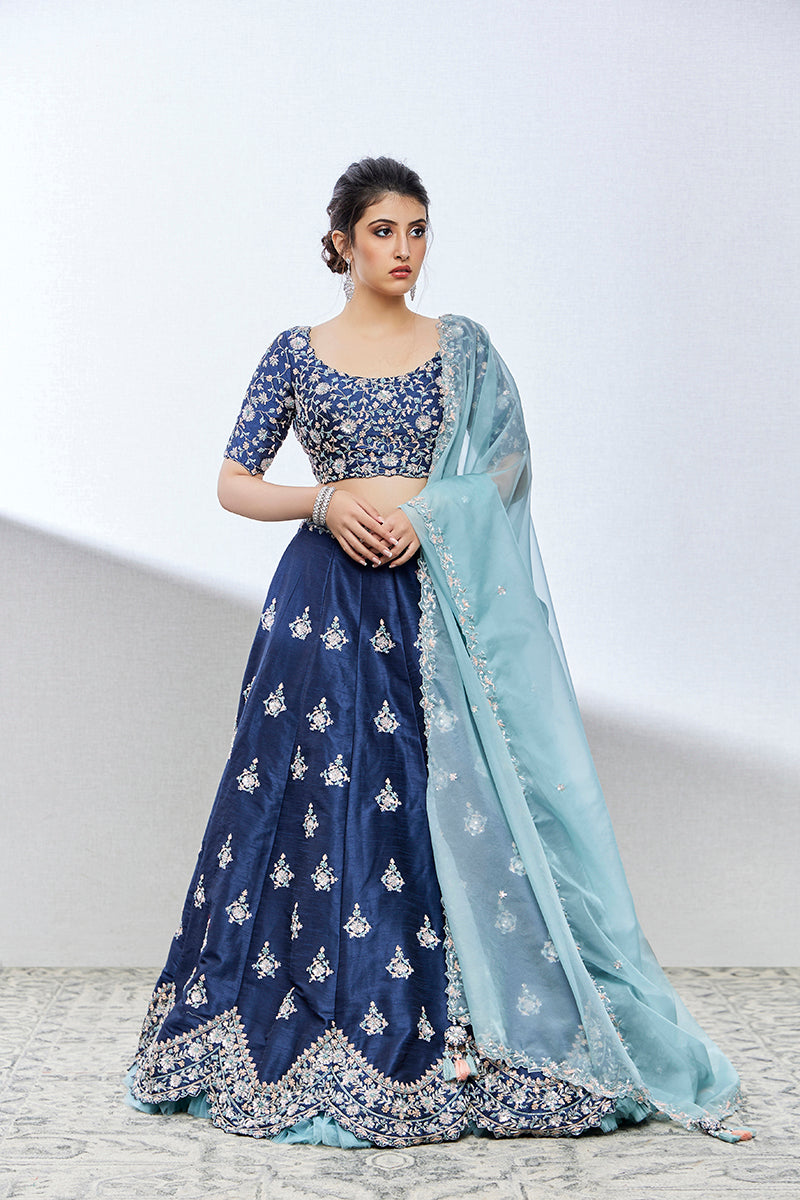 A woman poses in a greyish blue silk lehenga from BenzerWorld ethnicwear store, with her hands clasped in front of her waist.