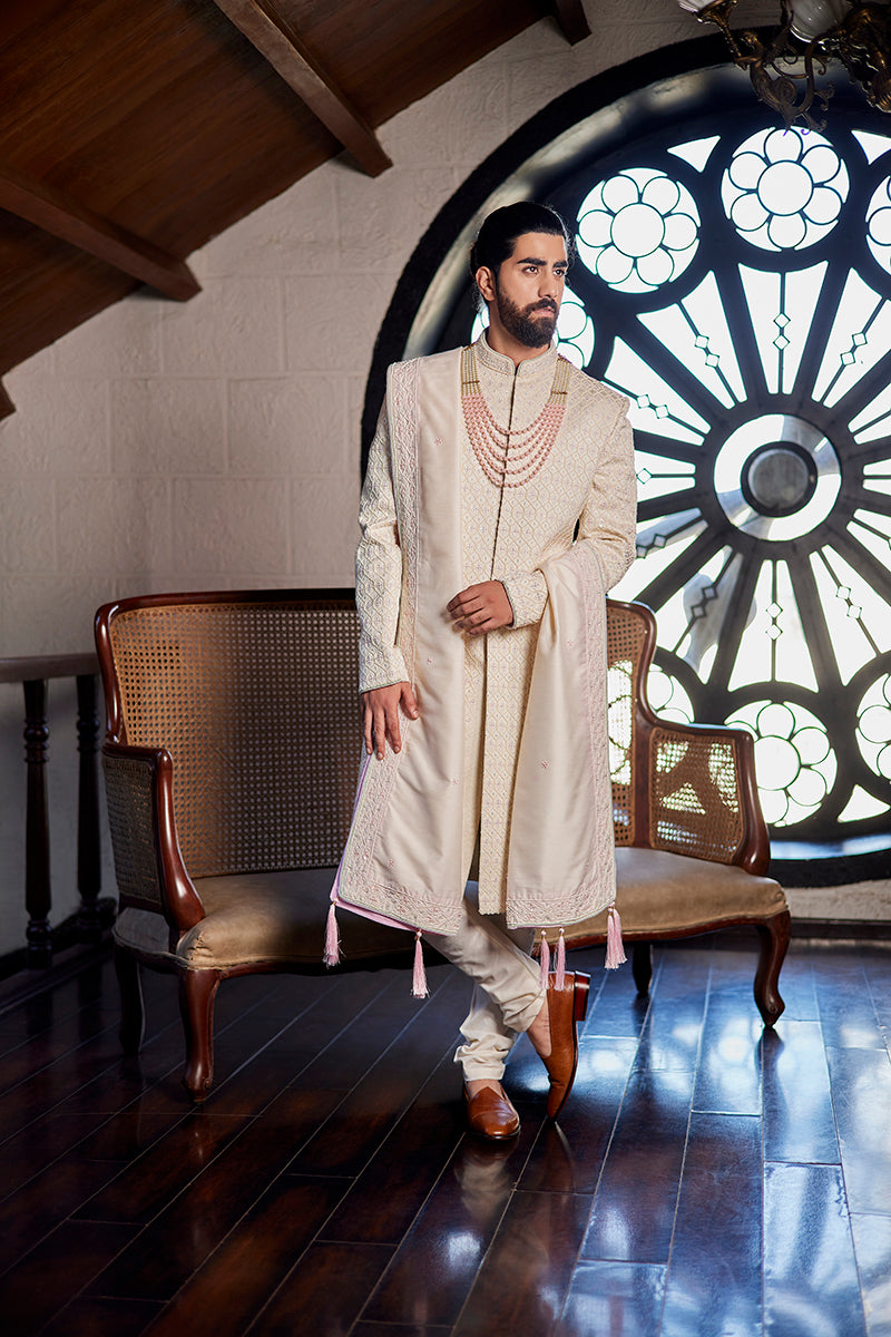 A man in an ivory brocade sherwani from BenzerWorld ethnicwear store poses in front of a pole.