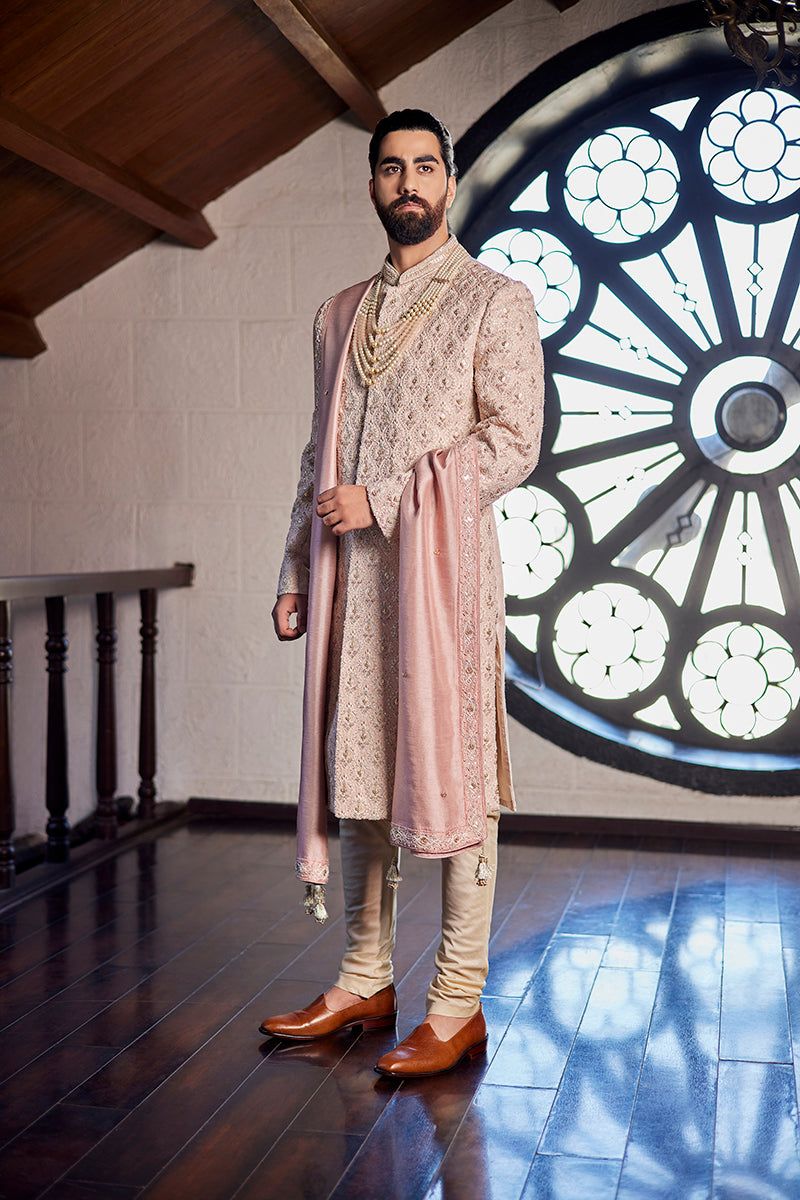 A man in a light pink brocade sherwani from BenzerWorld ethnicwear store poses in front of a window in an attic.