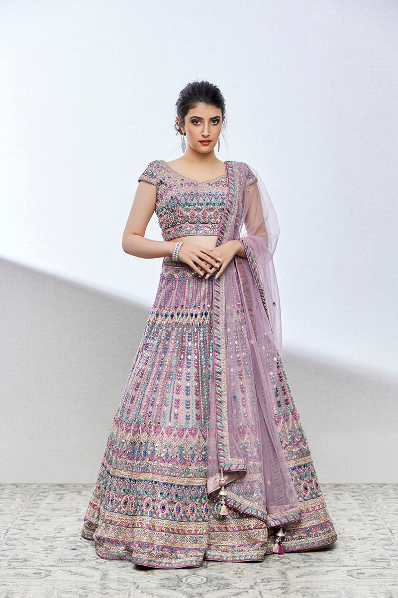 A woman poses in a light purple net lehenga from BenzerWorld ethnicwear store, with her hands clasped in front of her waist.