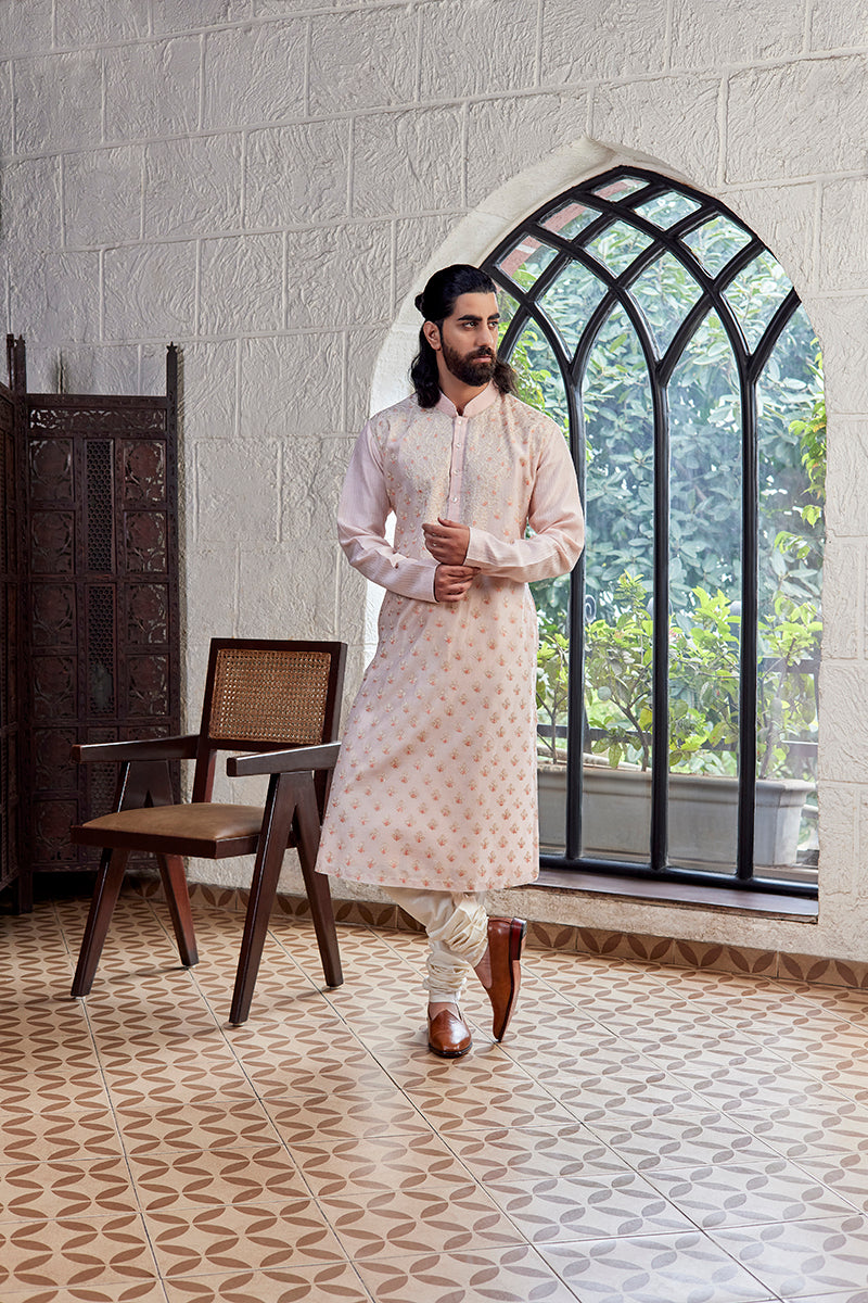 A man in a light pink chanderi silk kurta set from BenzerWorld ethnicwear store poses in front of an arched window.