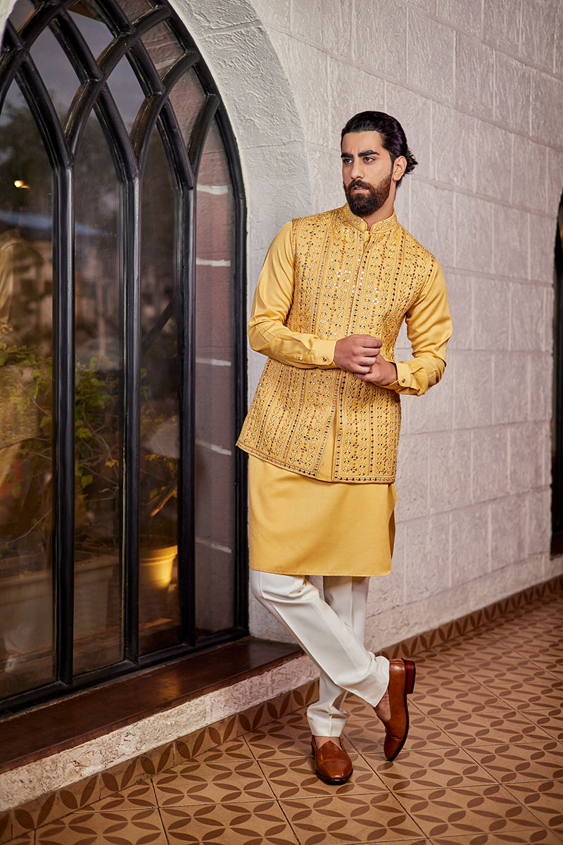 A man in a mustard poly-silk kurta and bundi set from BenzerWorld ethnicwear store poses in front of an arched window.