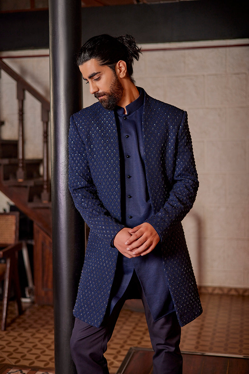 A man in a blue jacquard open-style Indo-Western suit from BenzerWorld ethnicwear store poses in front of a pole.